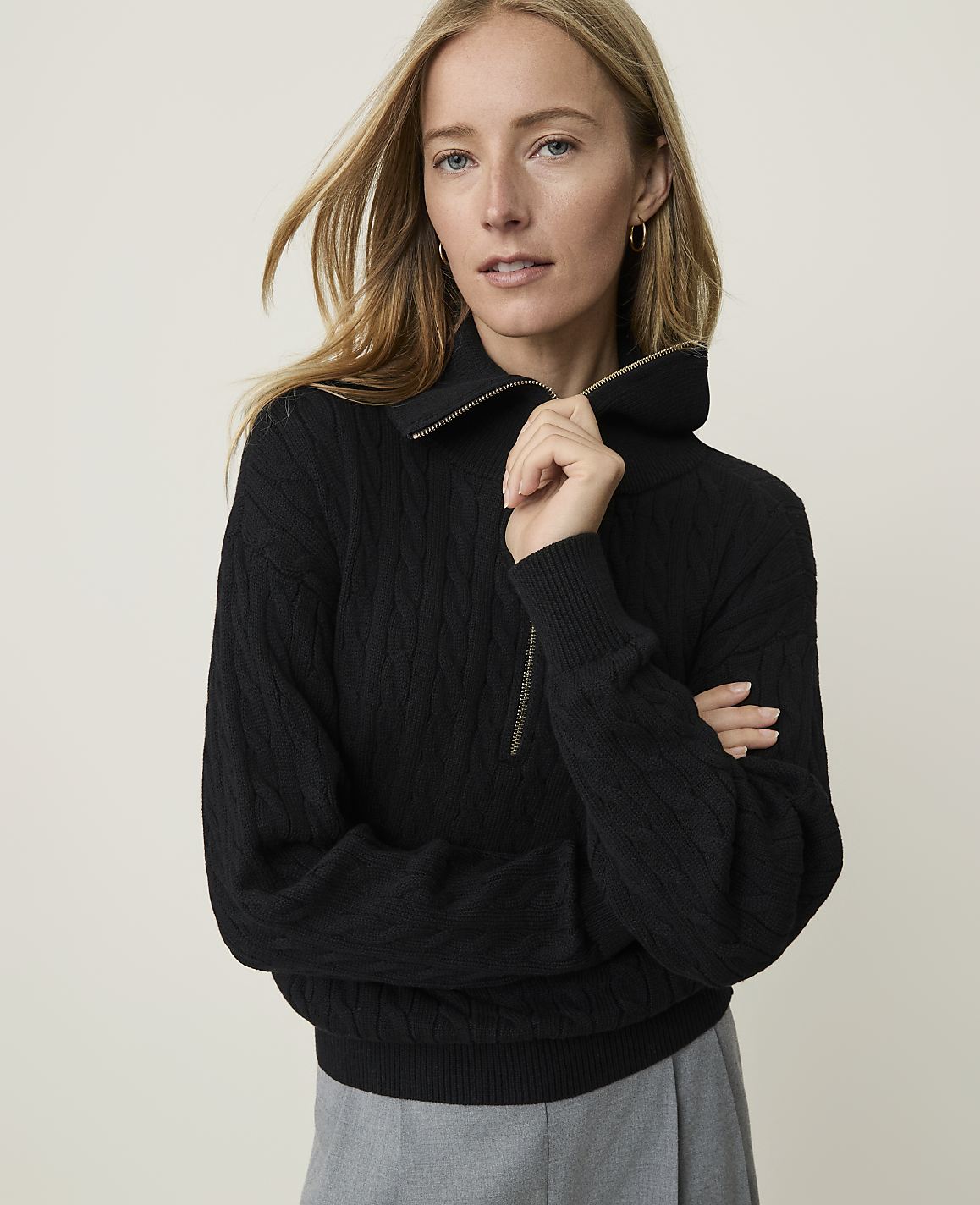 A woman in a black cable-knit sweater with a gold zipper stands against a plain, light background.