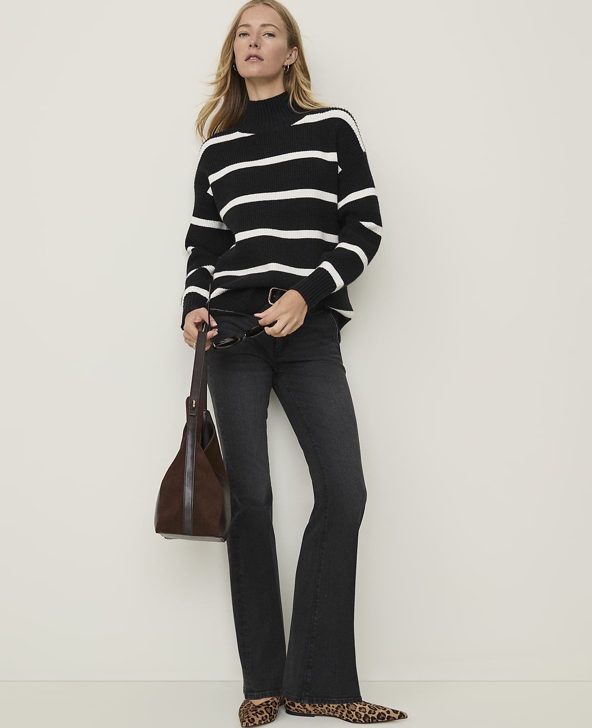 A woman in a black and white striped turtleneck sweater and dark flared jeans stands against a plain white background, holding a brown leather handbag and wearing leopard print flats.