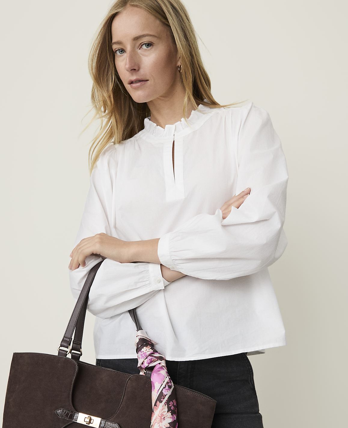 A woman in a white ruffled blouse holds a brown suede tote bag with a floral scarf in front of a plain, light-colored background.