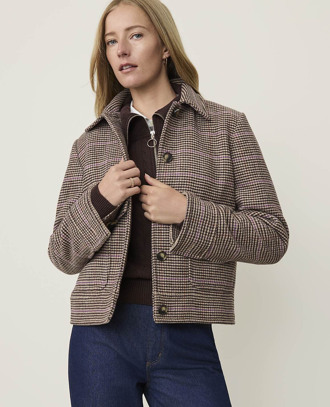 A woman in a plaid jacket and dark jeans stands against a plain, light-colored background.