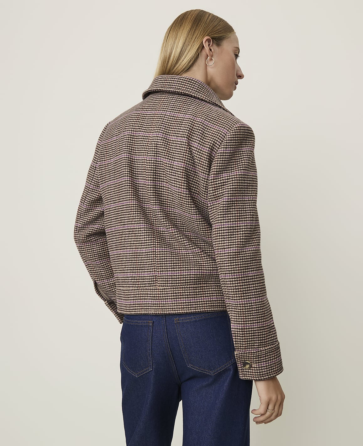 A woman in a brown and pink plaid jacket and dark jeans stands against a plain, light-colored background.