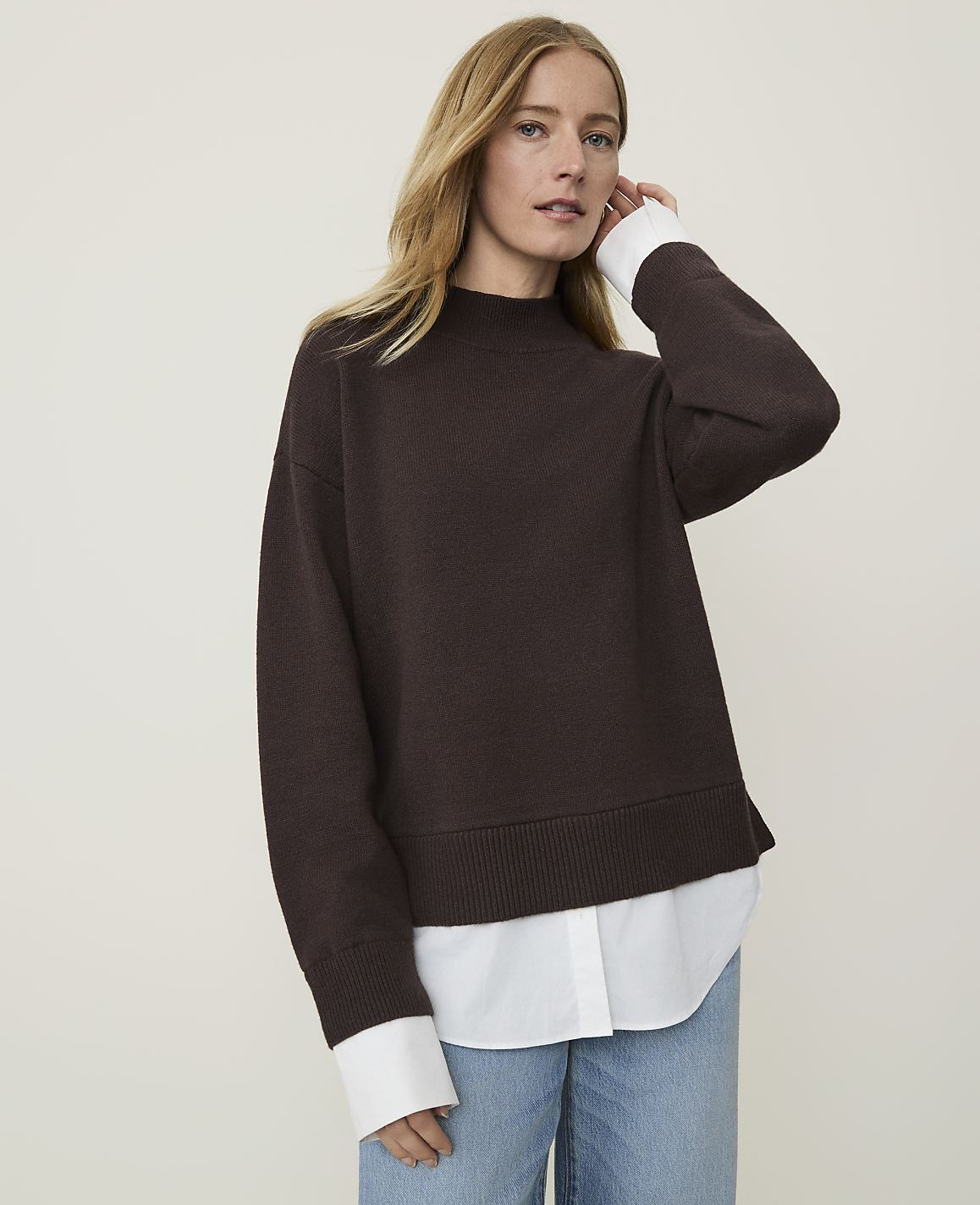 A woman in a brown sweater layered over a white shirt stands against a plain, light-colored background.
