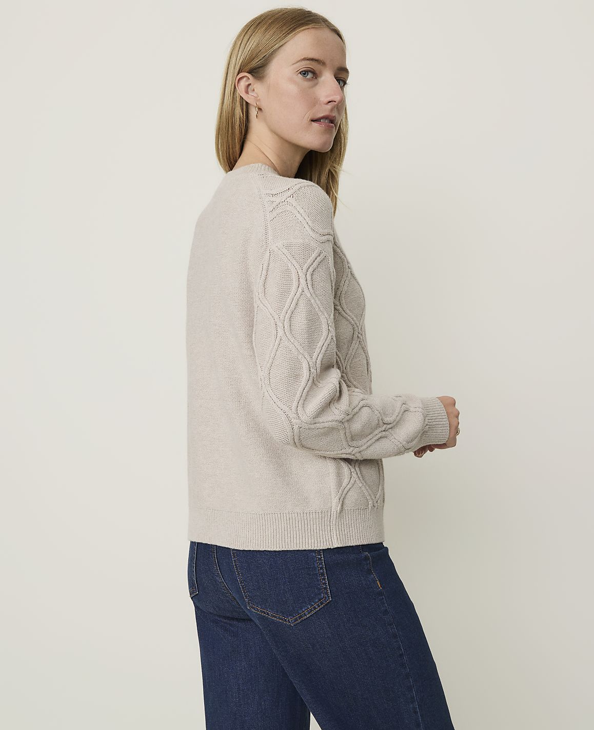 A woman in a beige cable-knit sweater and blue jeans stands against a plain, light-colored background.