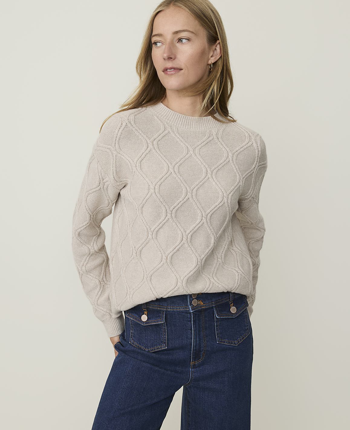 A woman in a beige cable-knit sweater and dark denim jeans stands against a plain, light-colored background.