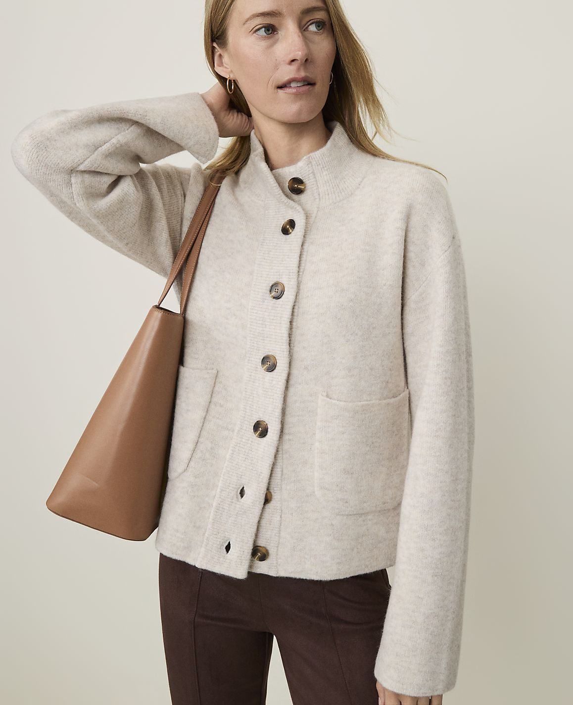 A woman in a cream cardigan and brown pants poses with a tan tote bag against a plain background.