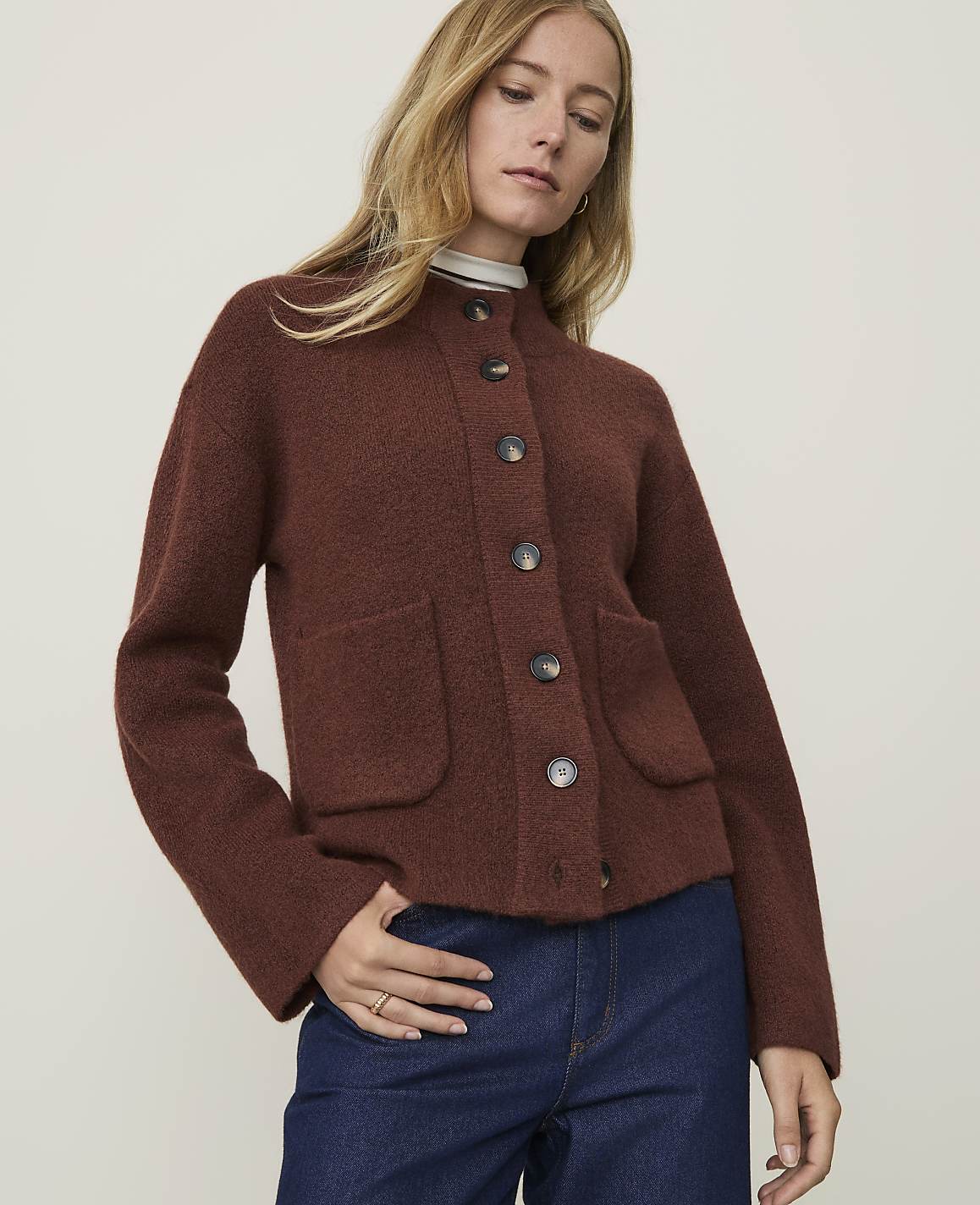 A woman in a brown cardigan and blue jeans stands against a plain, light-colored background.