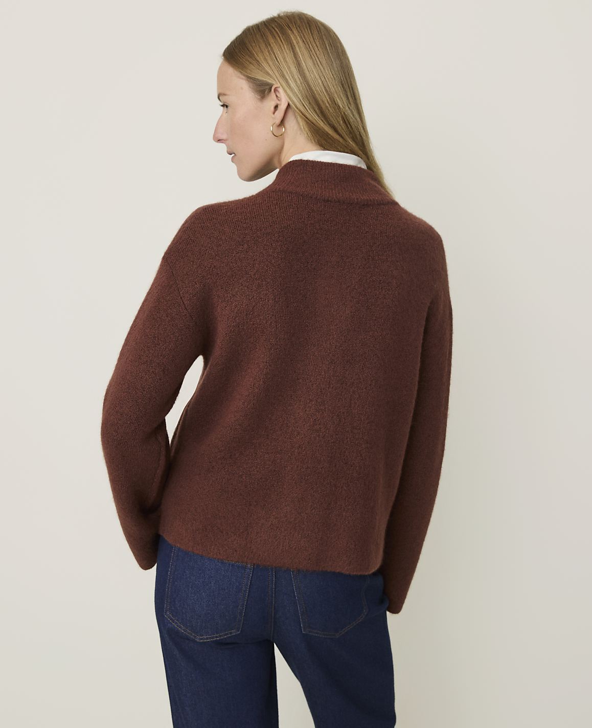 A woman in a brown sweater and blue jeans stands against a plain, light brown background.
