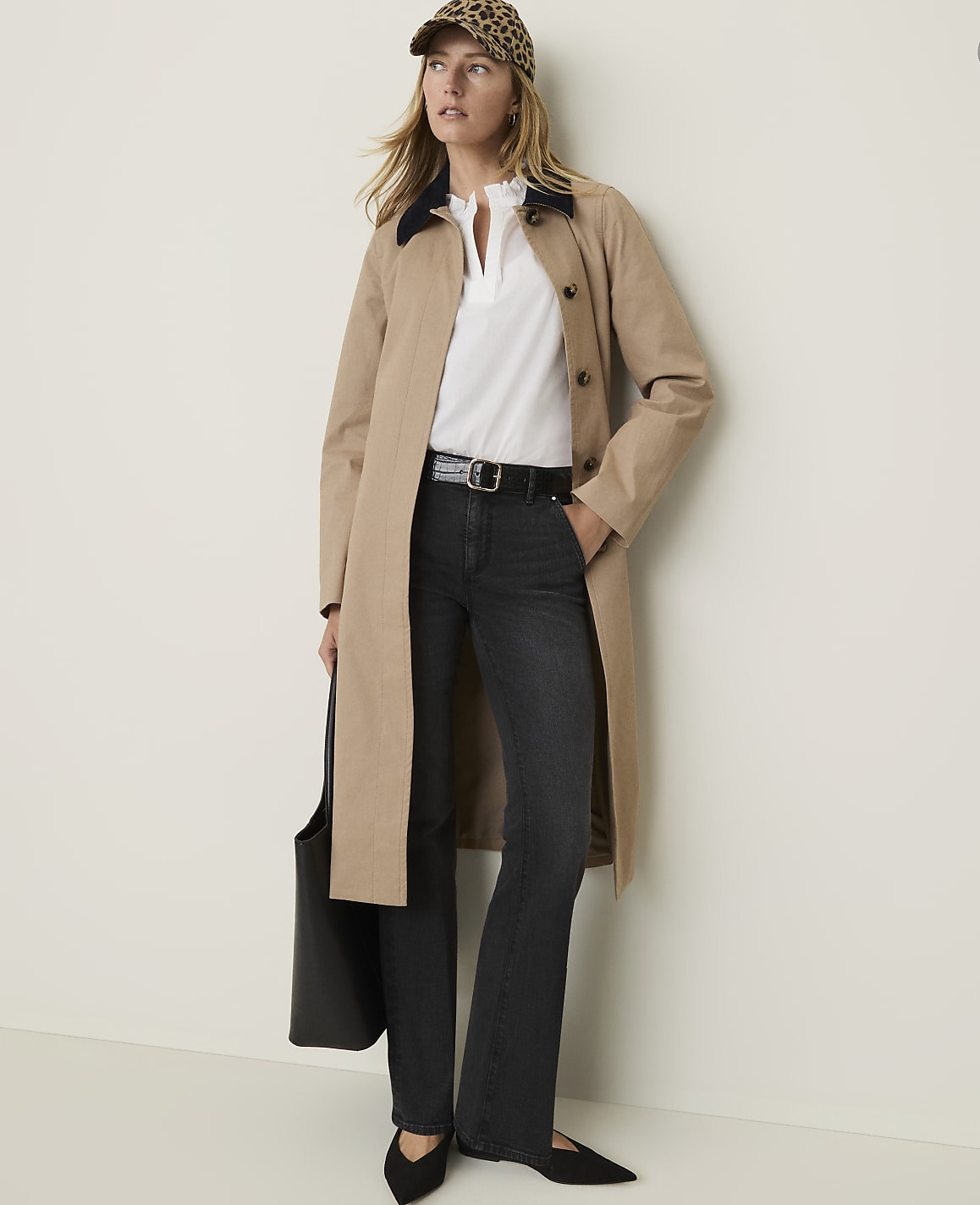 A woman in a tan trench coat, white blouse, dark jeans, and a leopard print cap stands against a plain, light-colored wall, holding a black tote bag.