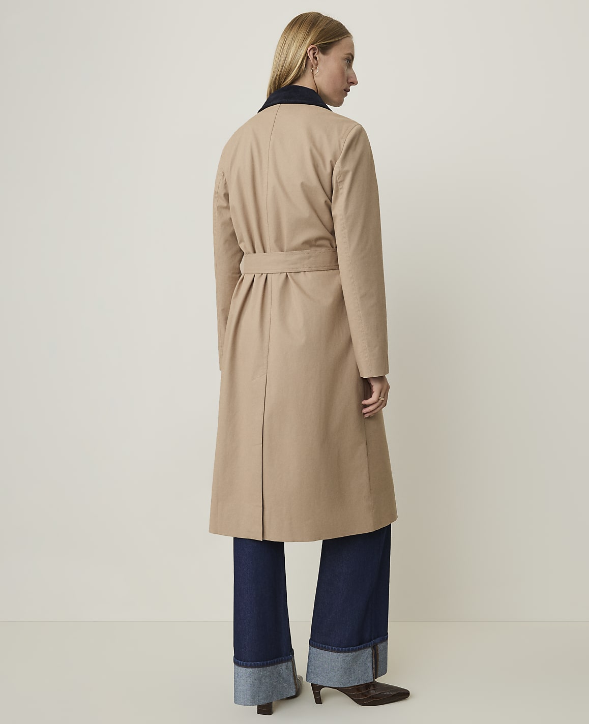 A woman in a tan trench coat and dark cuffed jeans stands against a plain, light background.