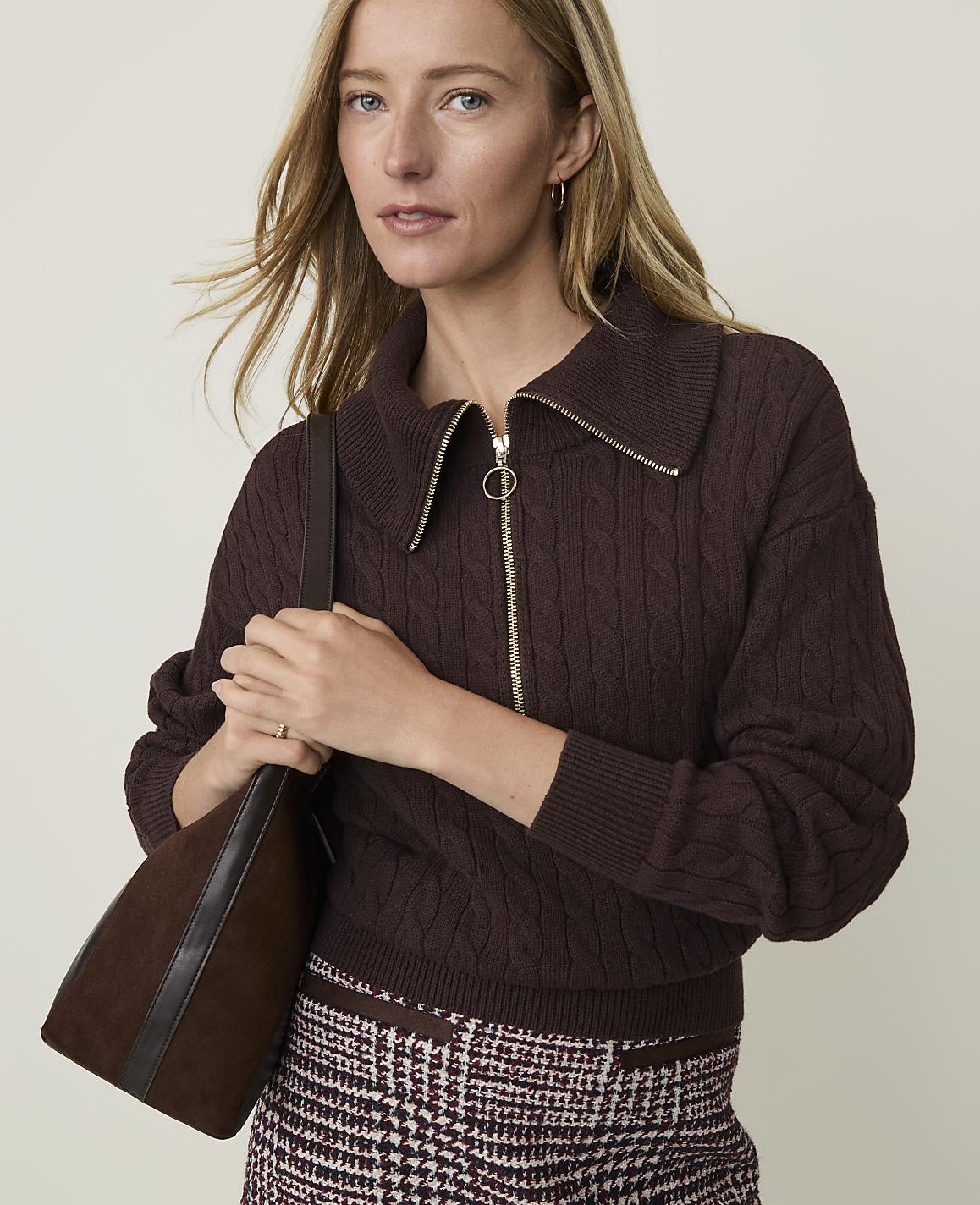 A woman in a brown cable-knit sweater and patterned skirt poses with a brown handbag against a plain background.