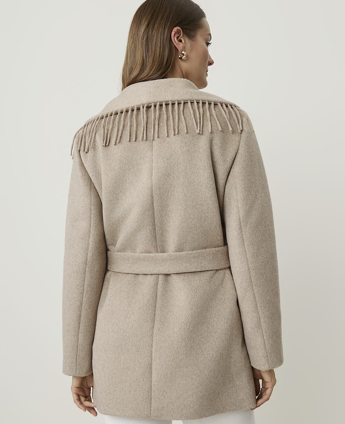A woman in a beige fringed coat stands against a plain, light background.