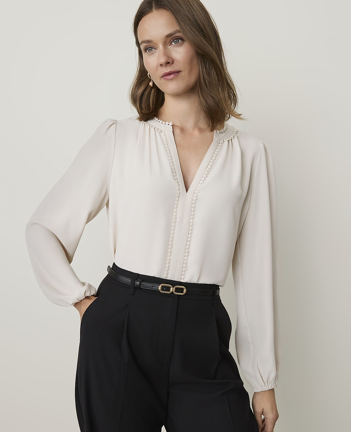 A woman in a cream blouse and black pants stands against a plain, light background.