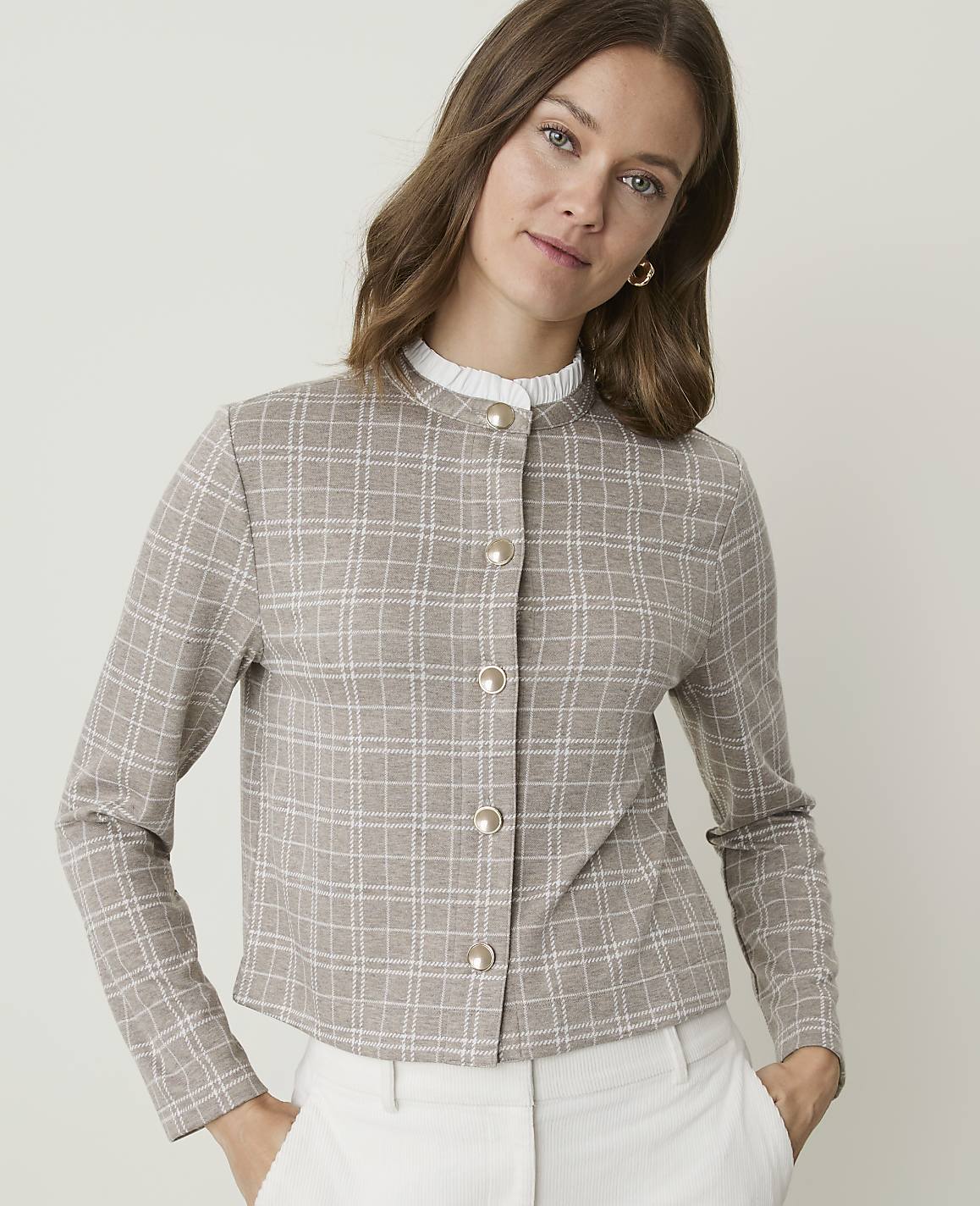 A woman in a plaid jacket and white pants stands against a plain background.