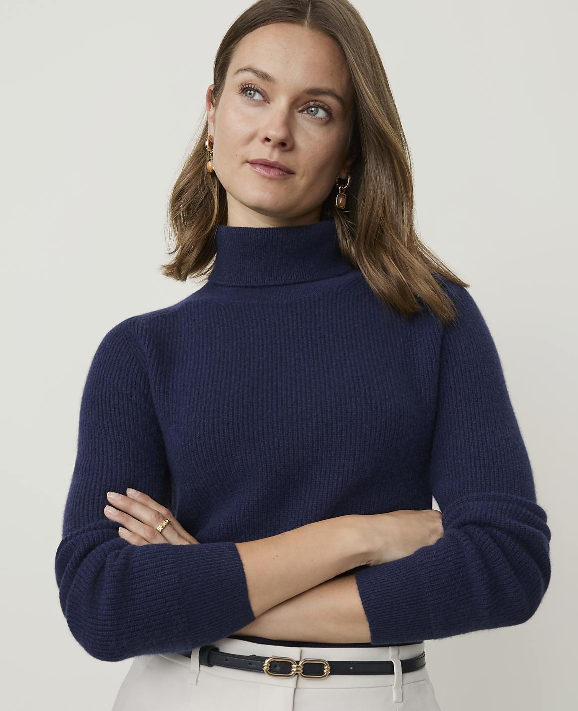 A woman in a navy turtleneck sweater and light-colored pants stands against a plain, light background.