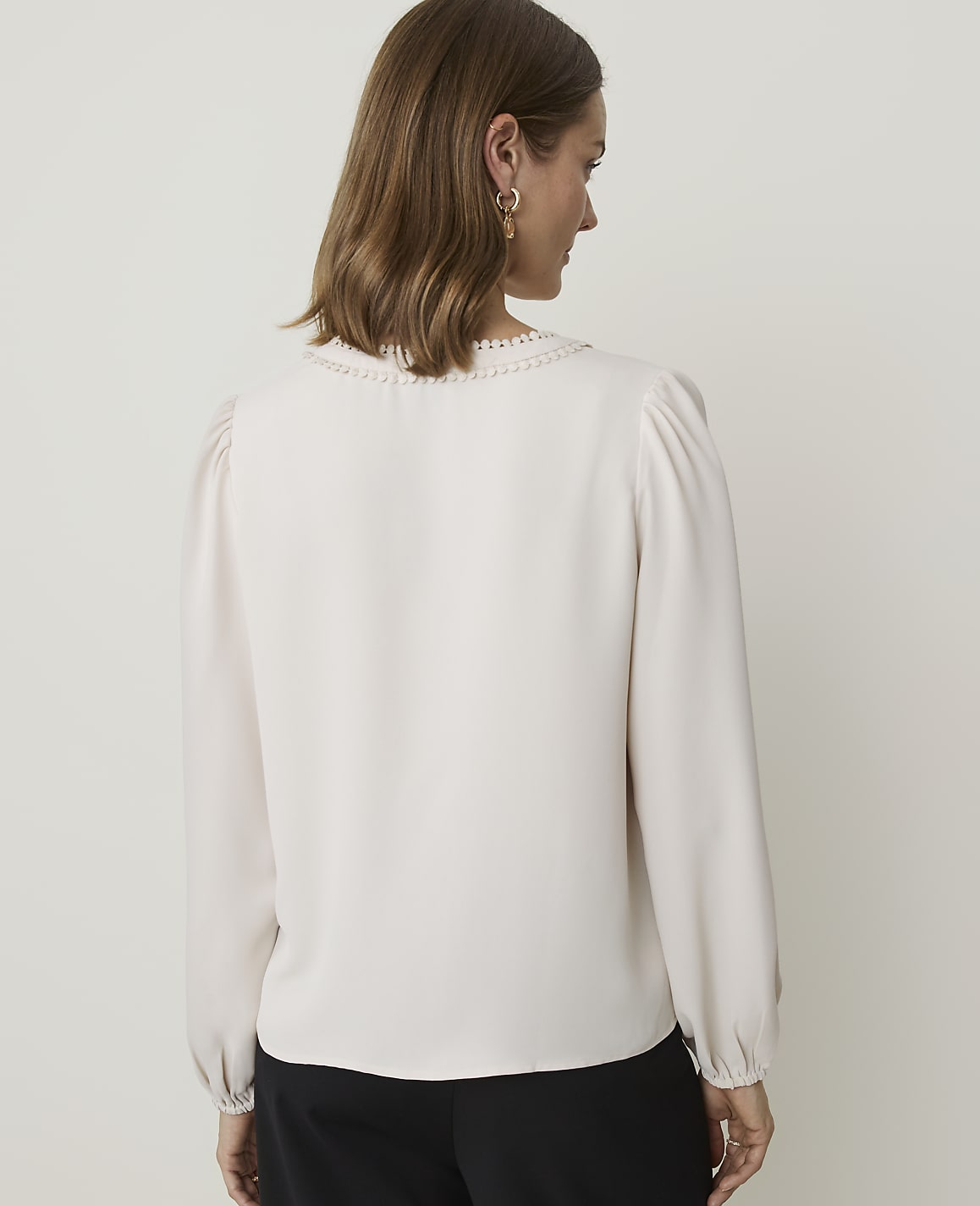 A woman in a cream-colored blouse with puffed sleeves and a decorative neckline stands against a plain, light gray background.