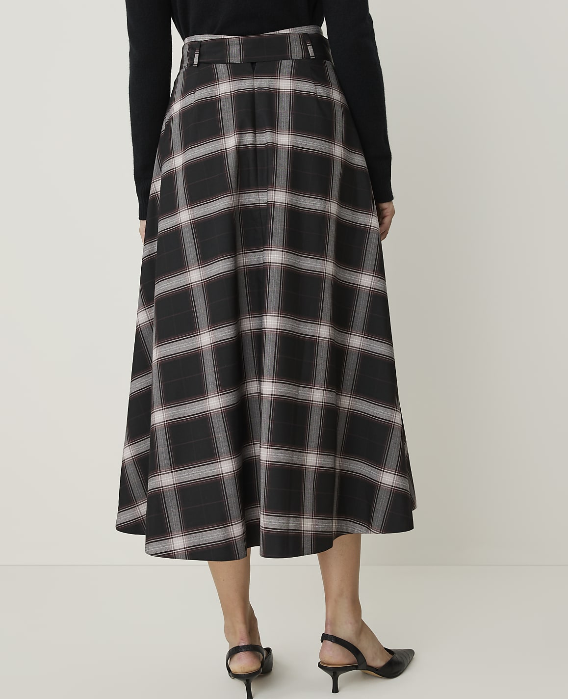 A woman in a black sweater and a plaid midi skirt stands against a plain white background.