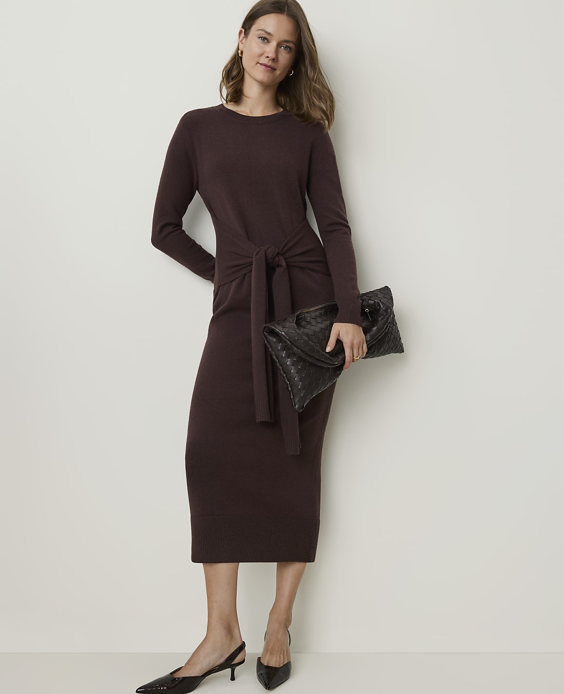A woman in a brown knit dress and black slingback heels holds a woven clutch in front of a plain white background.