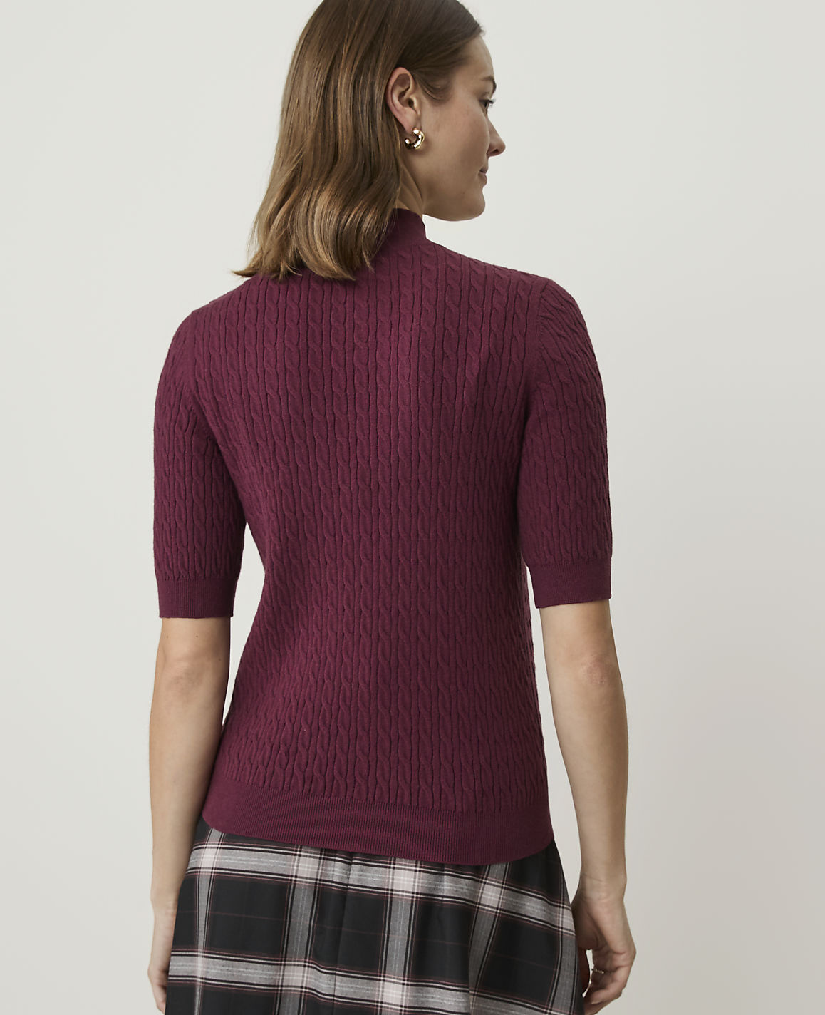A woman in a burgundy cable-knit turtleneck sweater and plaid skirt stands against a plain, light-colored background.