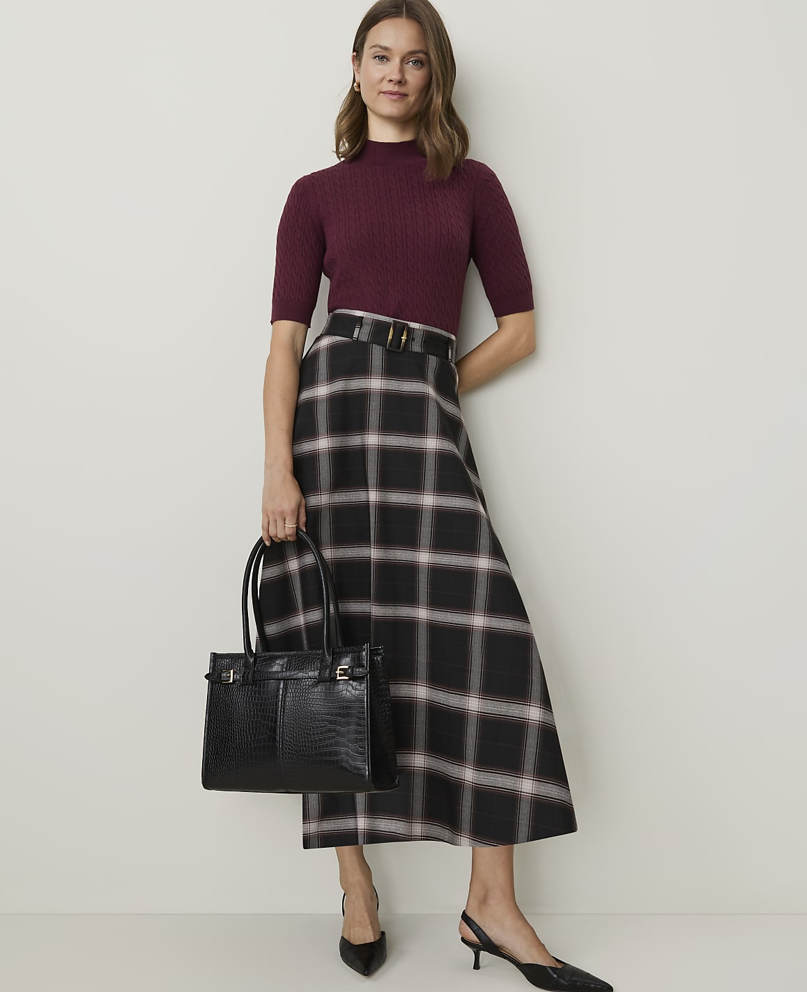 A woman in a burgundy mock-neck top and a plaid skirt stands holding a black crocodile-embossed tote bag against a plain, light-colored wall.