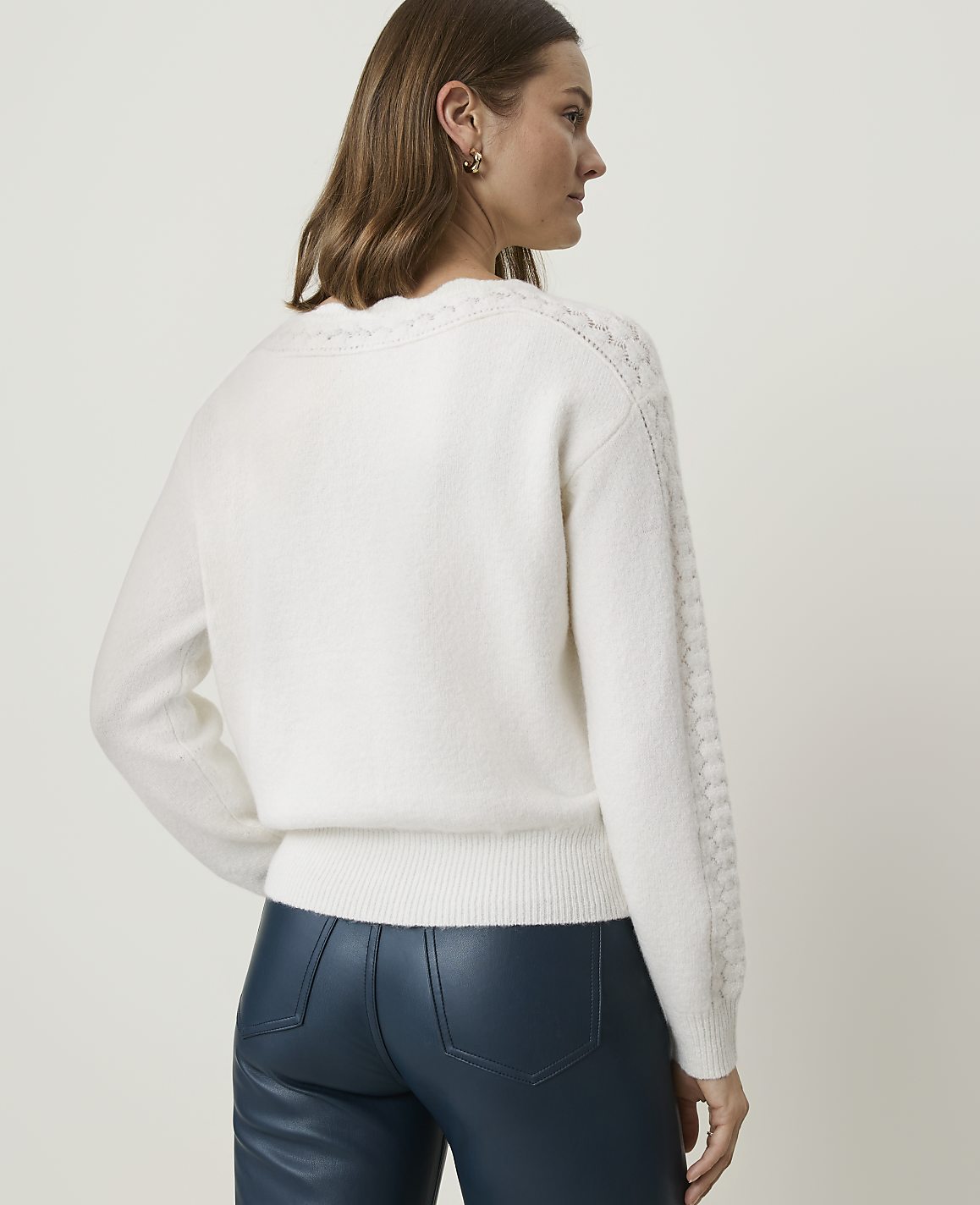 A woman in a white cable-knit sweater and blue leather pants stands against a plain white background.