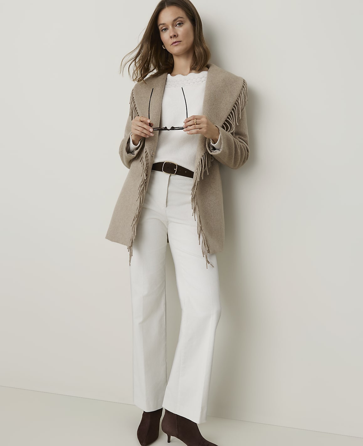 A woman in a fringed jacket and white pants stands against a plain, light-colored wall.