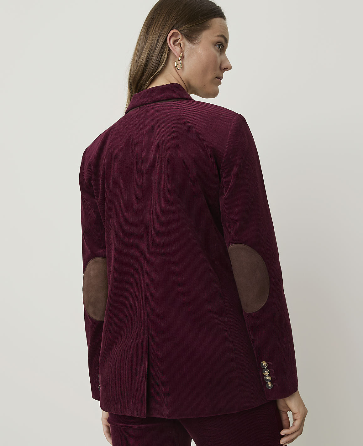 A woman in a burgundy corduroy blazer with suede elbow patches stands against a plain, light-colored background.