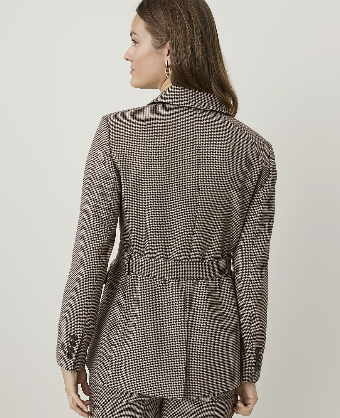 A woman in a brown checkered blazer and matching pants stands against a plain, light-colored background.