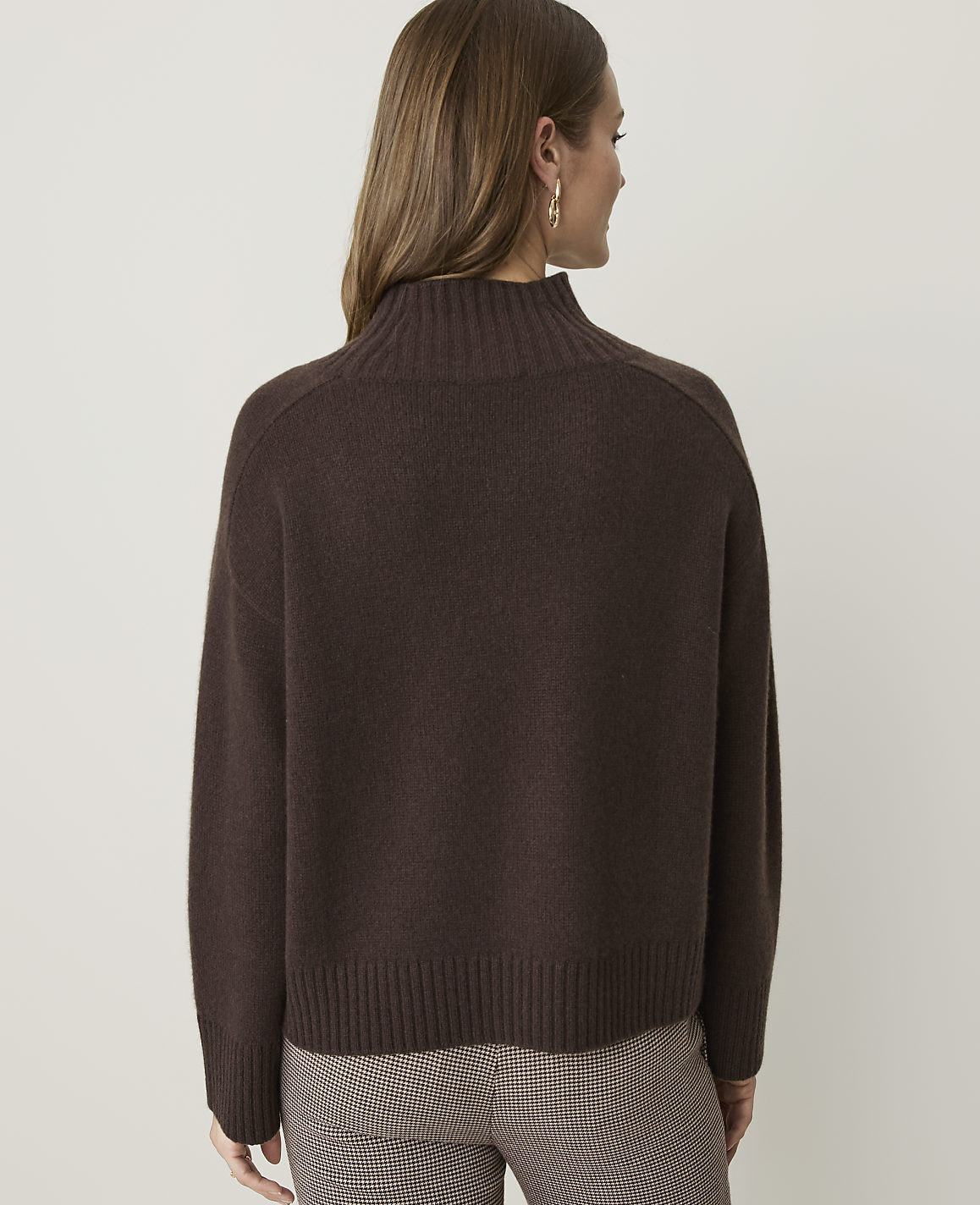A woman in a brown turtleneck sweater and plaid pants stands against a plain, light gray background.