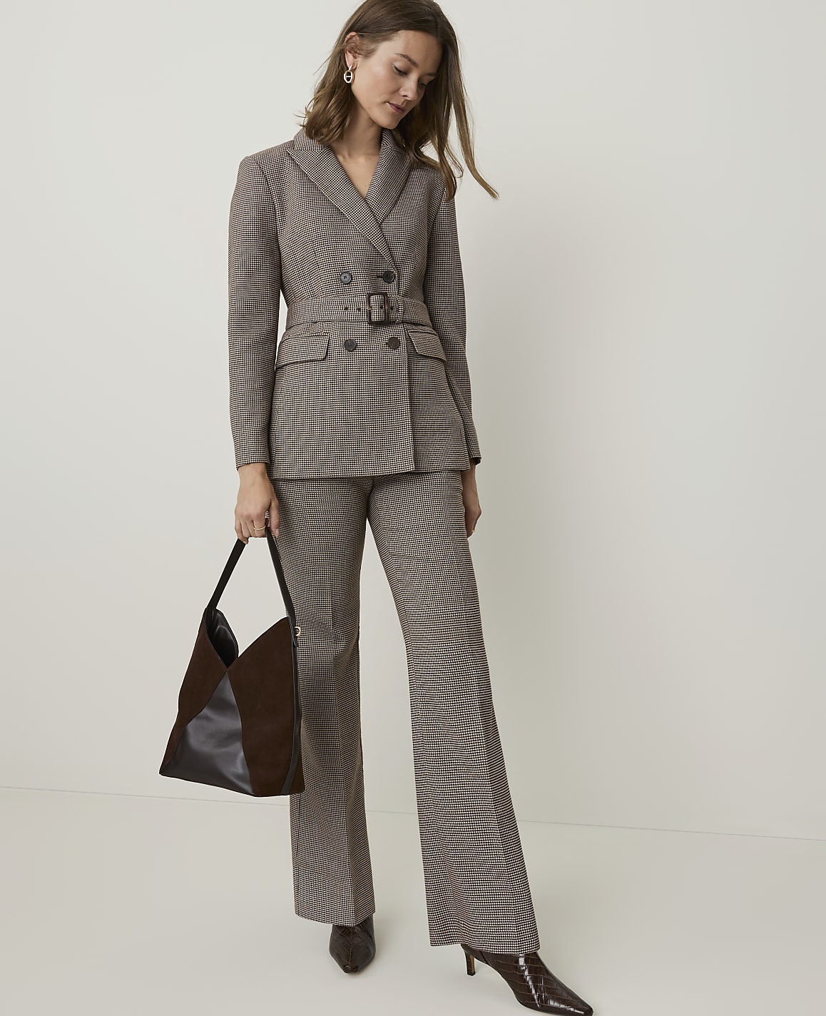 A woman in a brown checkered suit and dark brown boots stands with a dark brown bag in front of a plain white background.