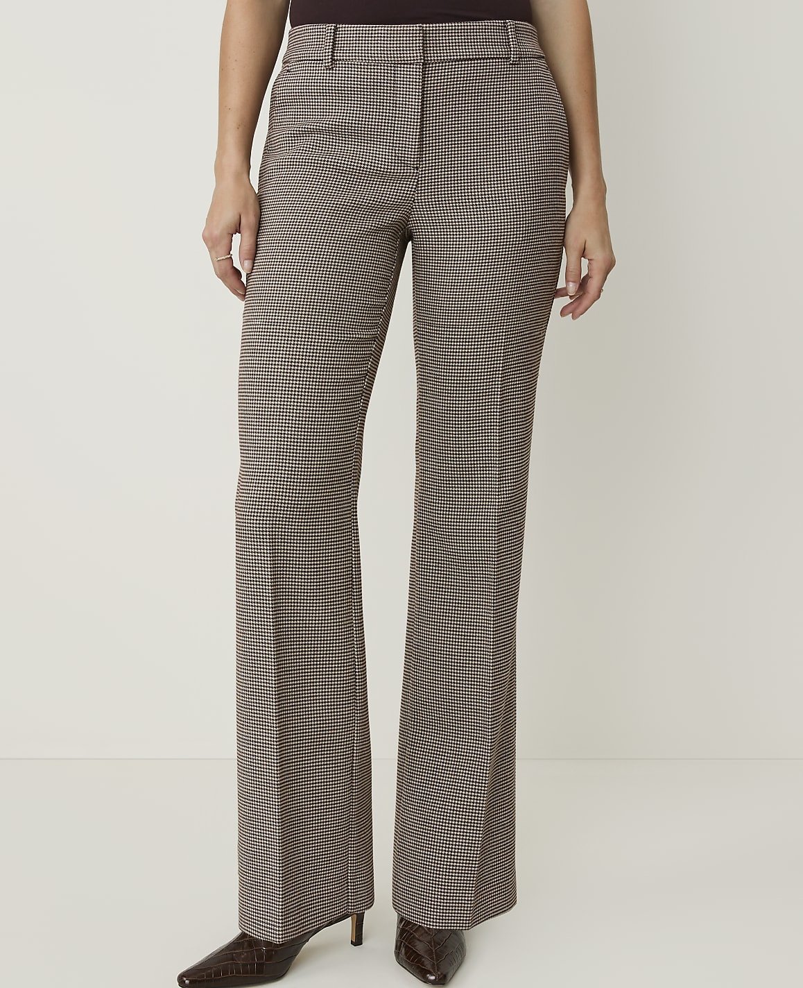 A person stands in front of a plain white background, wearing brown checkered flared pants and dark pointed-toe heels.