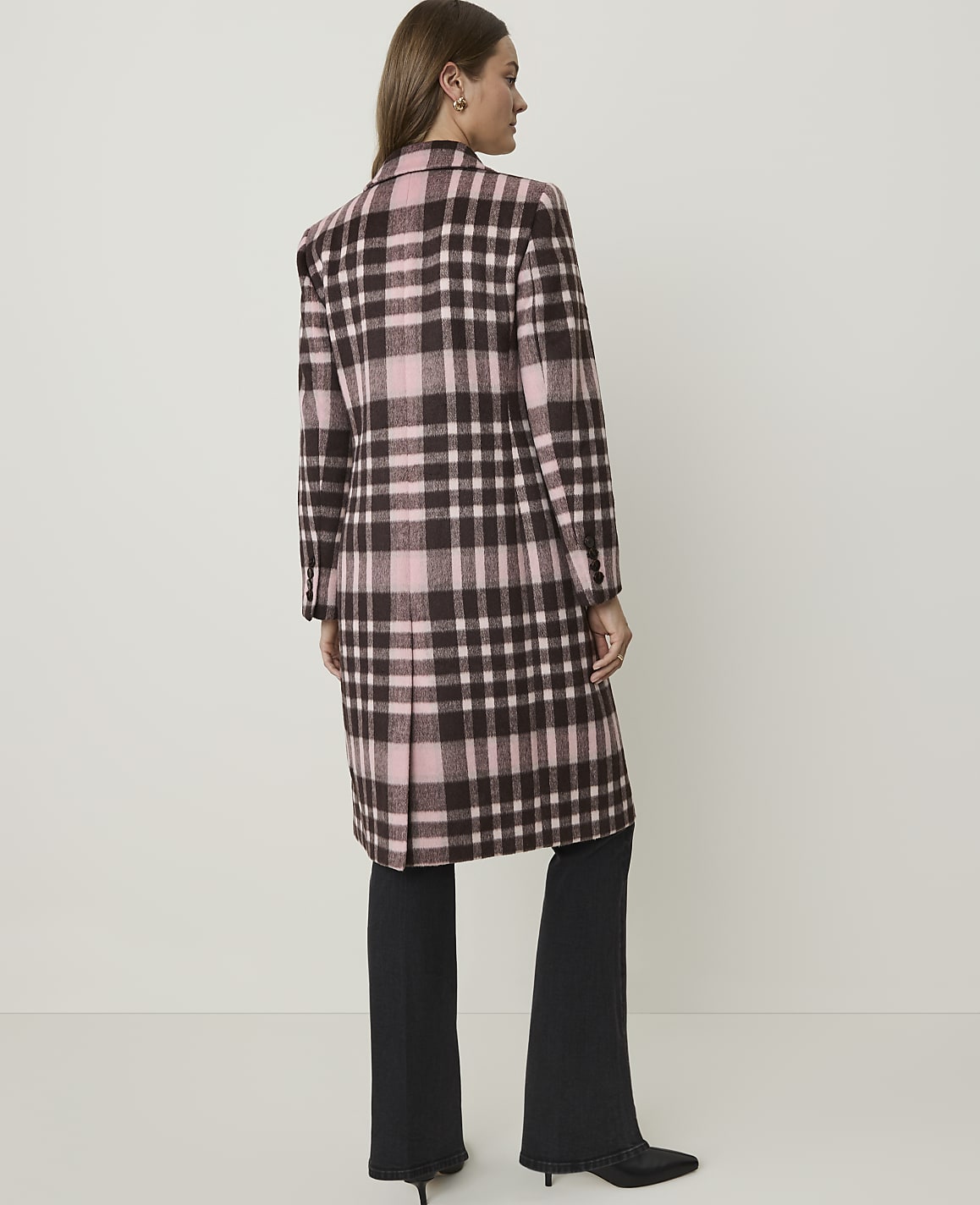A woman in a pink and brown plaid coat stands against a plain, light-colored background.
