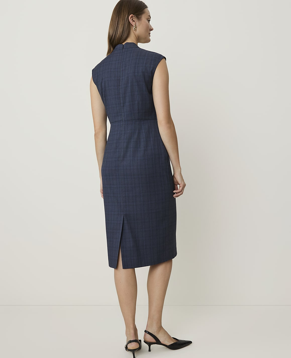 A woman in a blue plaid dress stands against a plain, light-colored background.