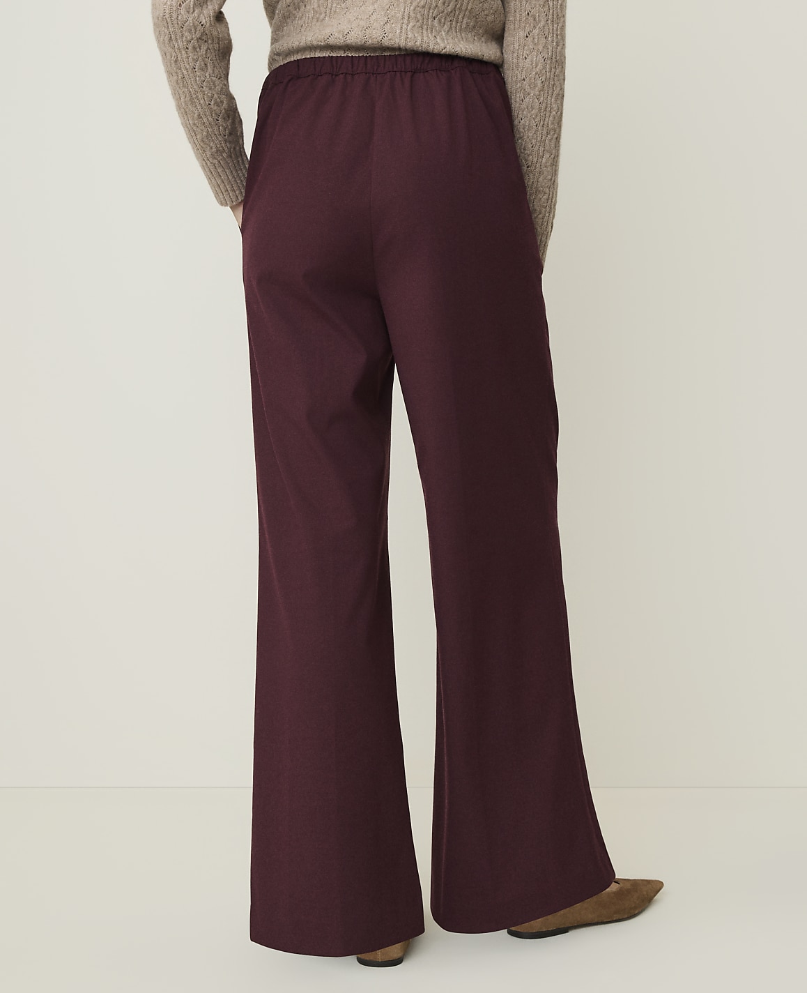 A person wearing burgundy flared pants and a beige sweater stands against a plain, light-colored background.