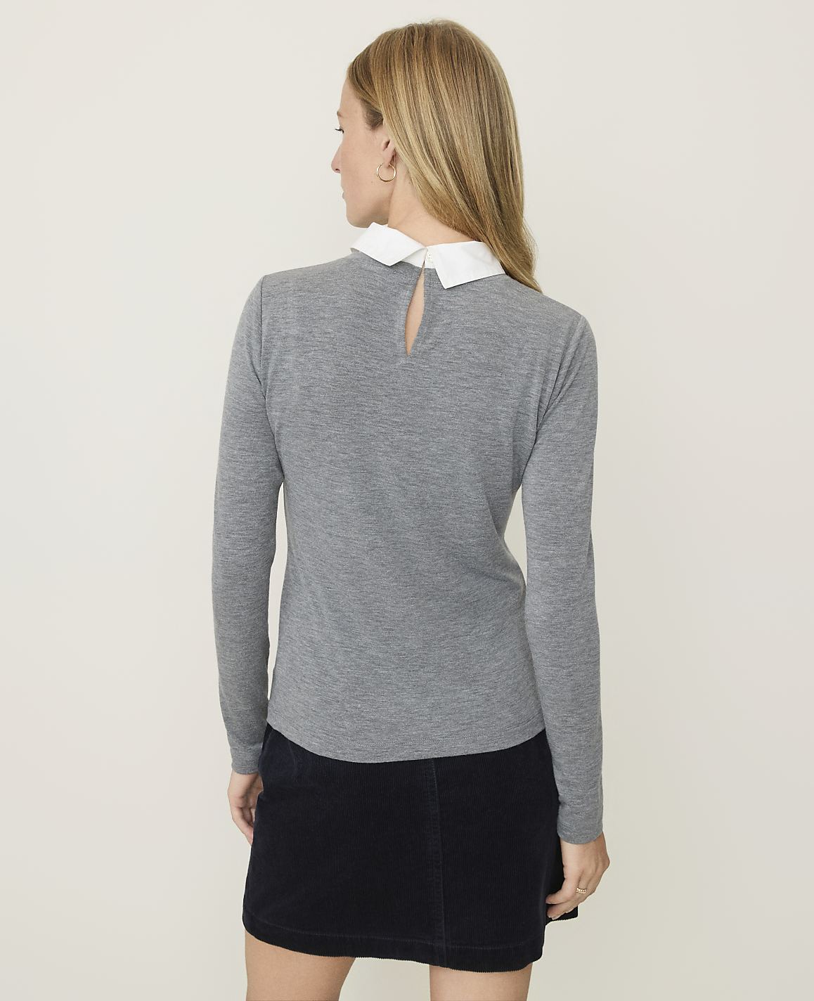 A woman in a gray sweater with a white collar and a dark skirt stands against a plain, light-colored background.
