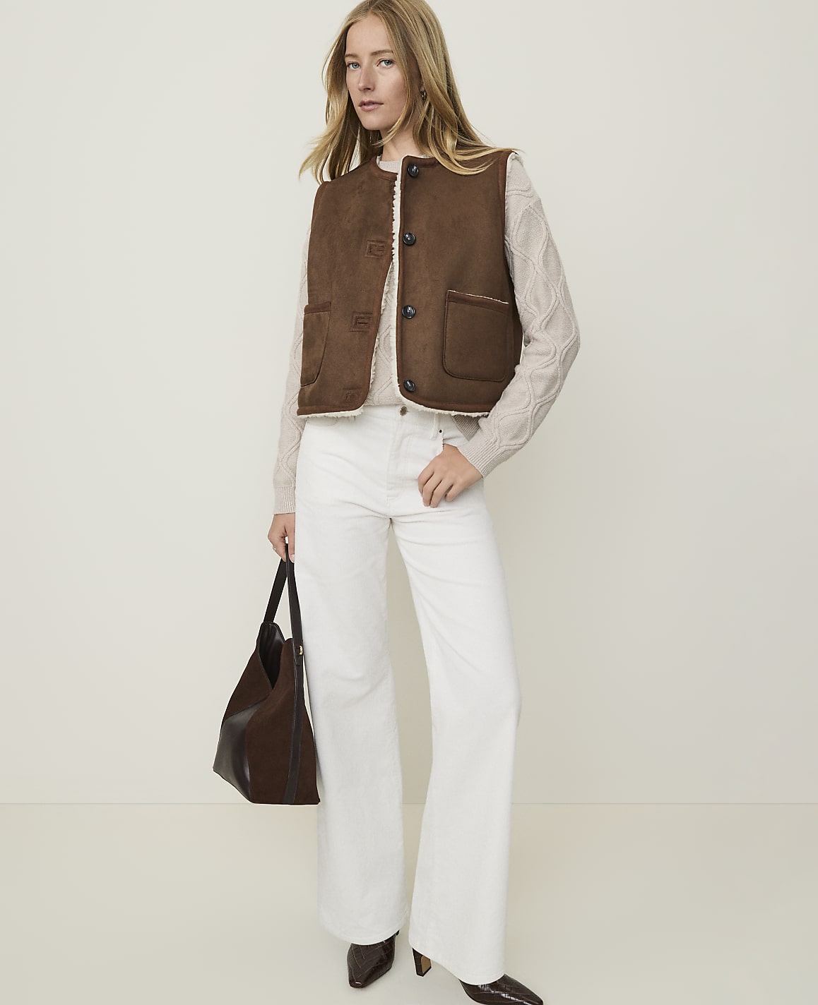 A woman in a brown vest and white pants stands against a plain, light background, holding a dark brown purse.