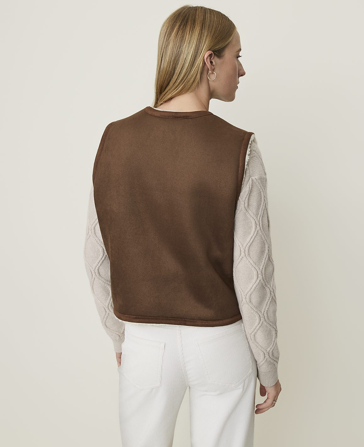 A woman in a brown vest and cream sweater stands against a plain, light-colored background.