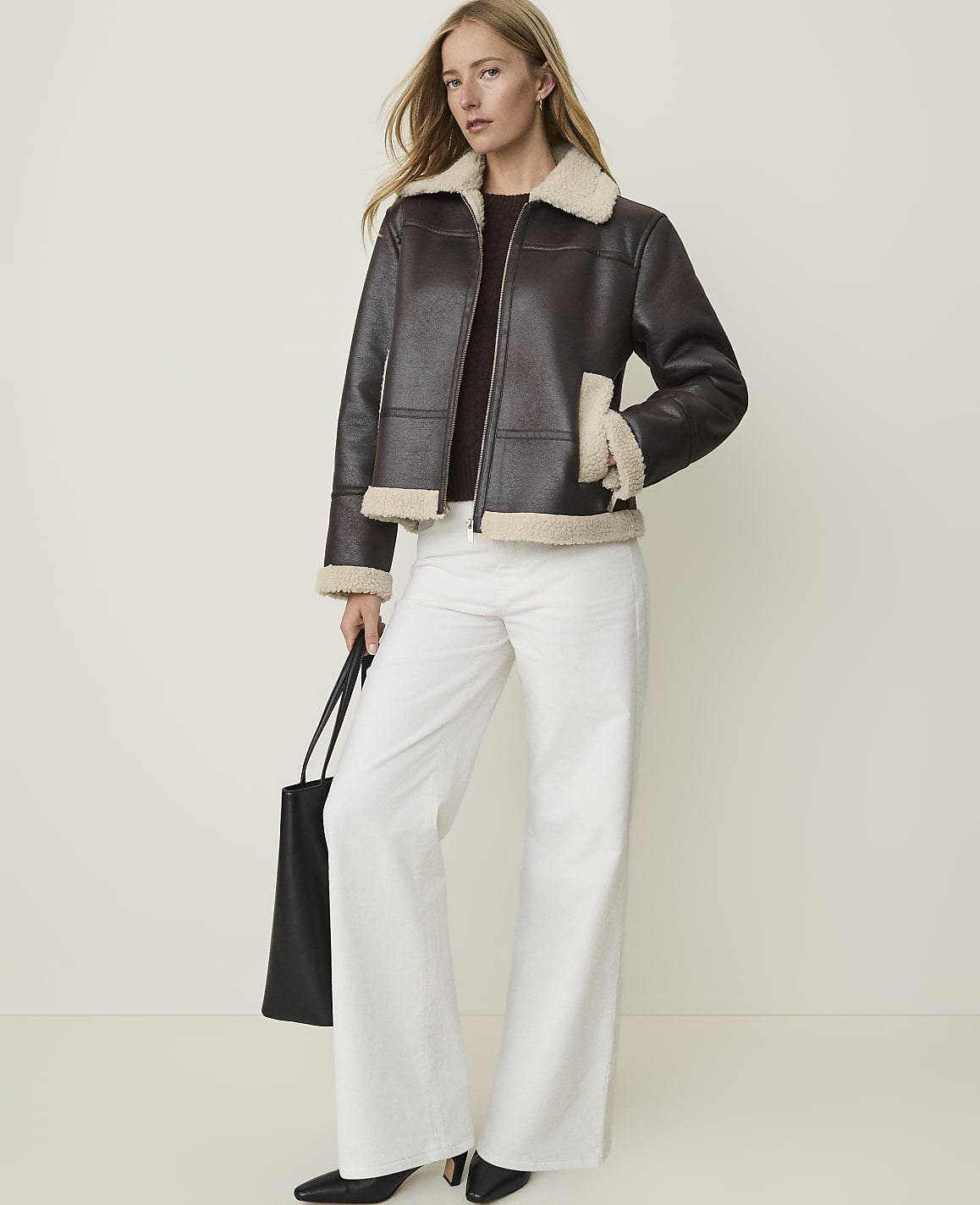 A woman in a brown shearling-lined jacket and white pants stands against a plain, light background, holding a black tote bag.