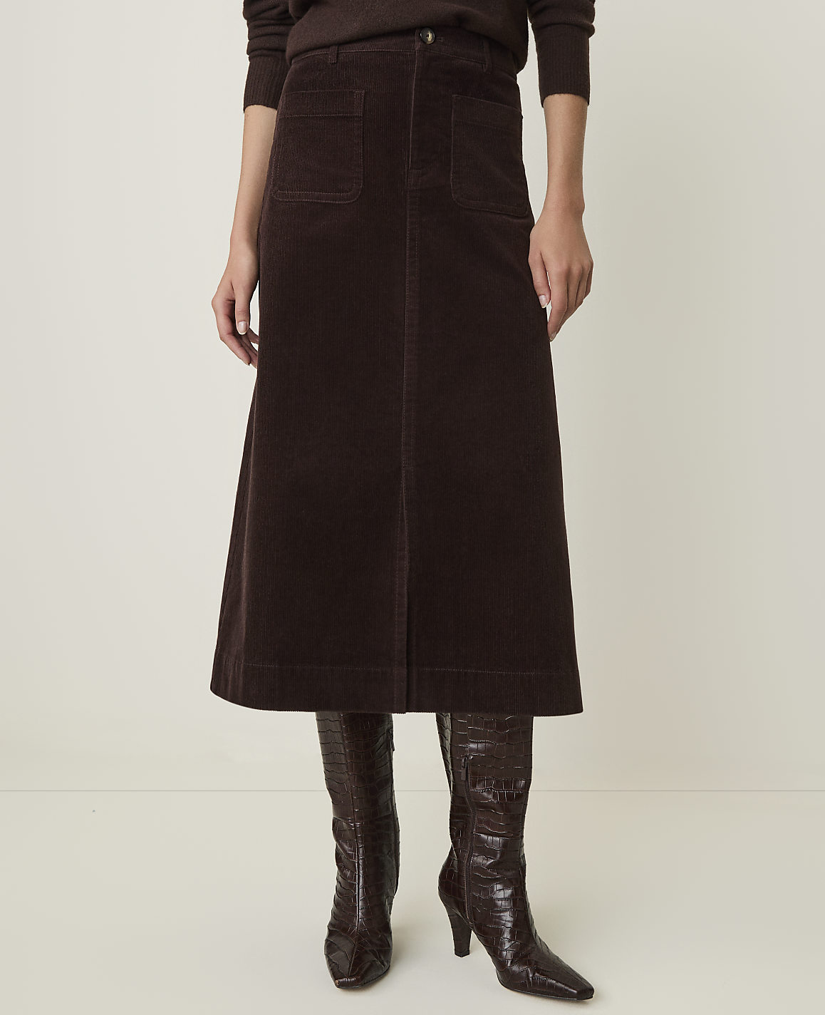 A person stands in a brown corduroy skirt and croc-embossed boots against a plain, light-colored background.
