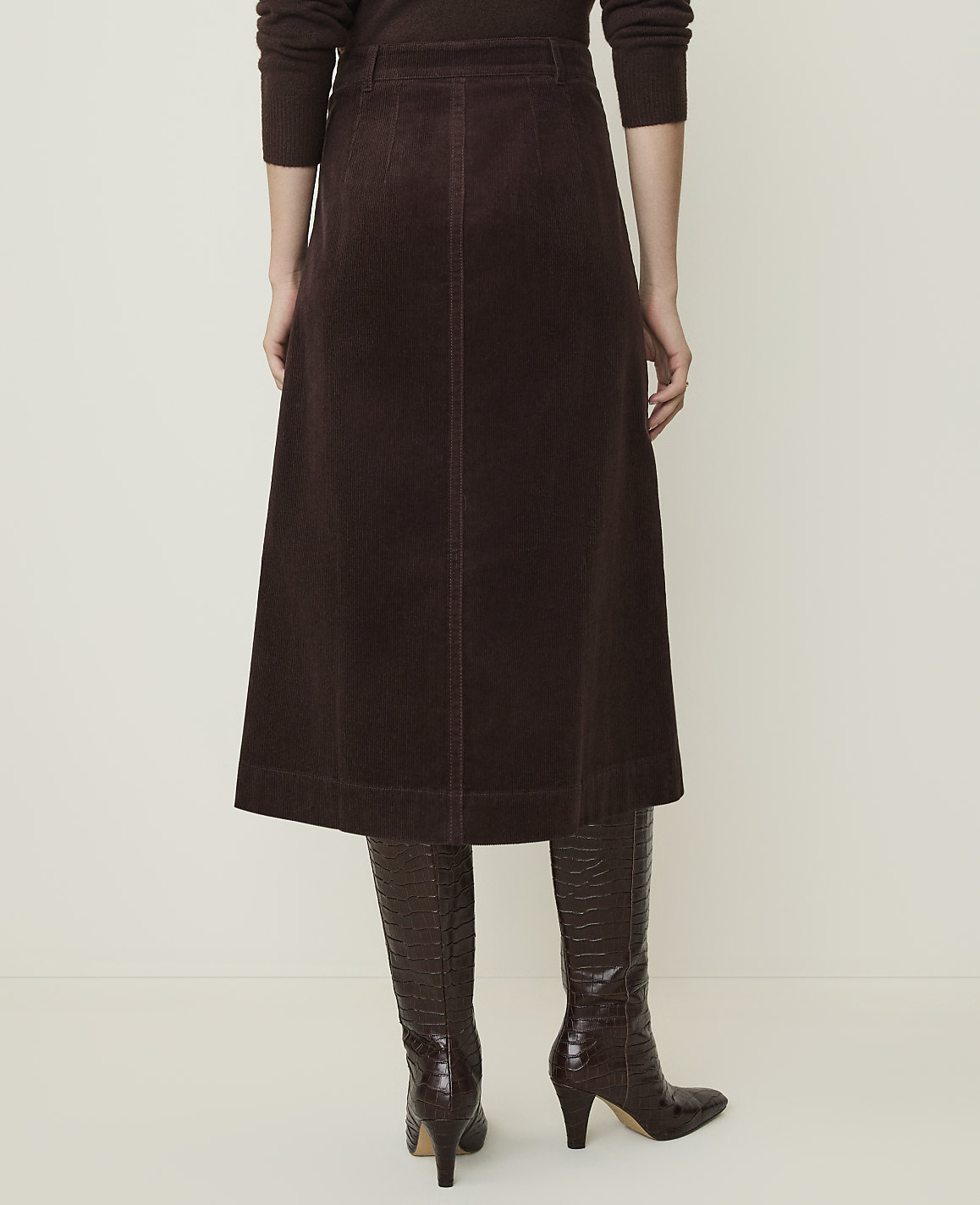 A person wearing a brown corduroy skirt and matching boots stands against a plain, light-colored background.