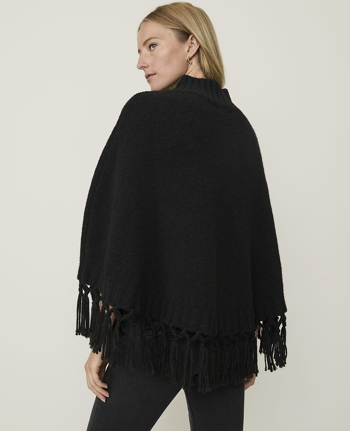 A woman in a black fringed poncho stands against a plain, light-colored background.