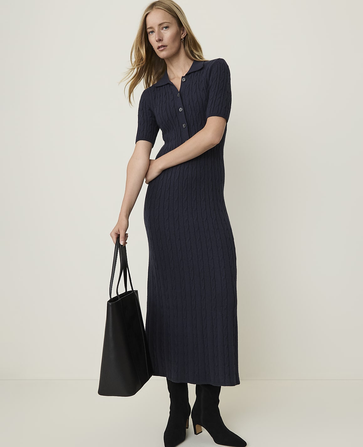 A woman in a navy cable-knit midi dress and black boots holds a black tote bag against a plain, light background.