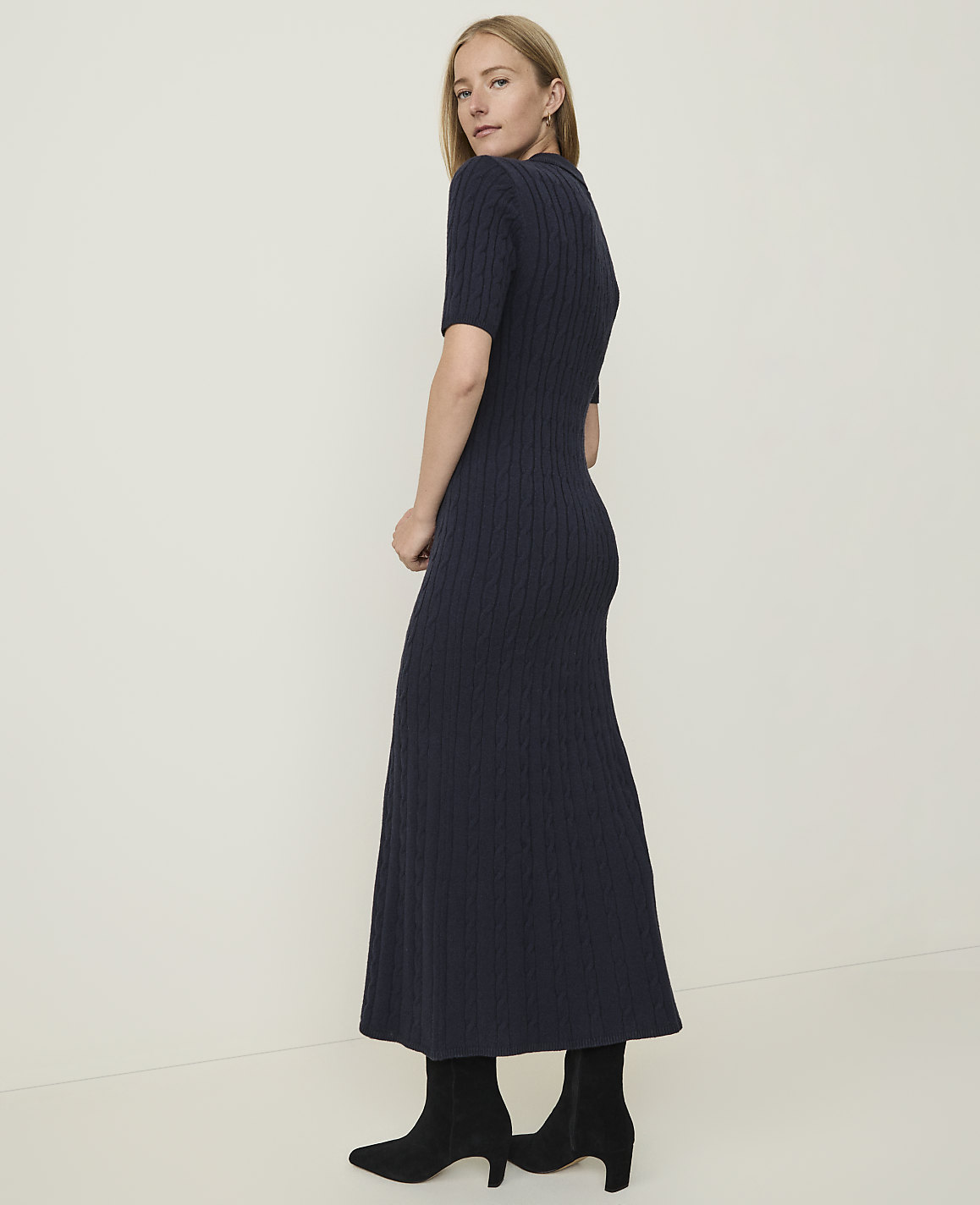 A woman in a navy cable-knit dress and black boots stands against a plain, light-colored background.