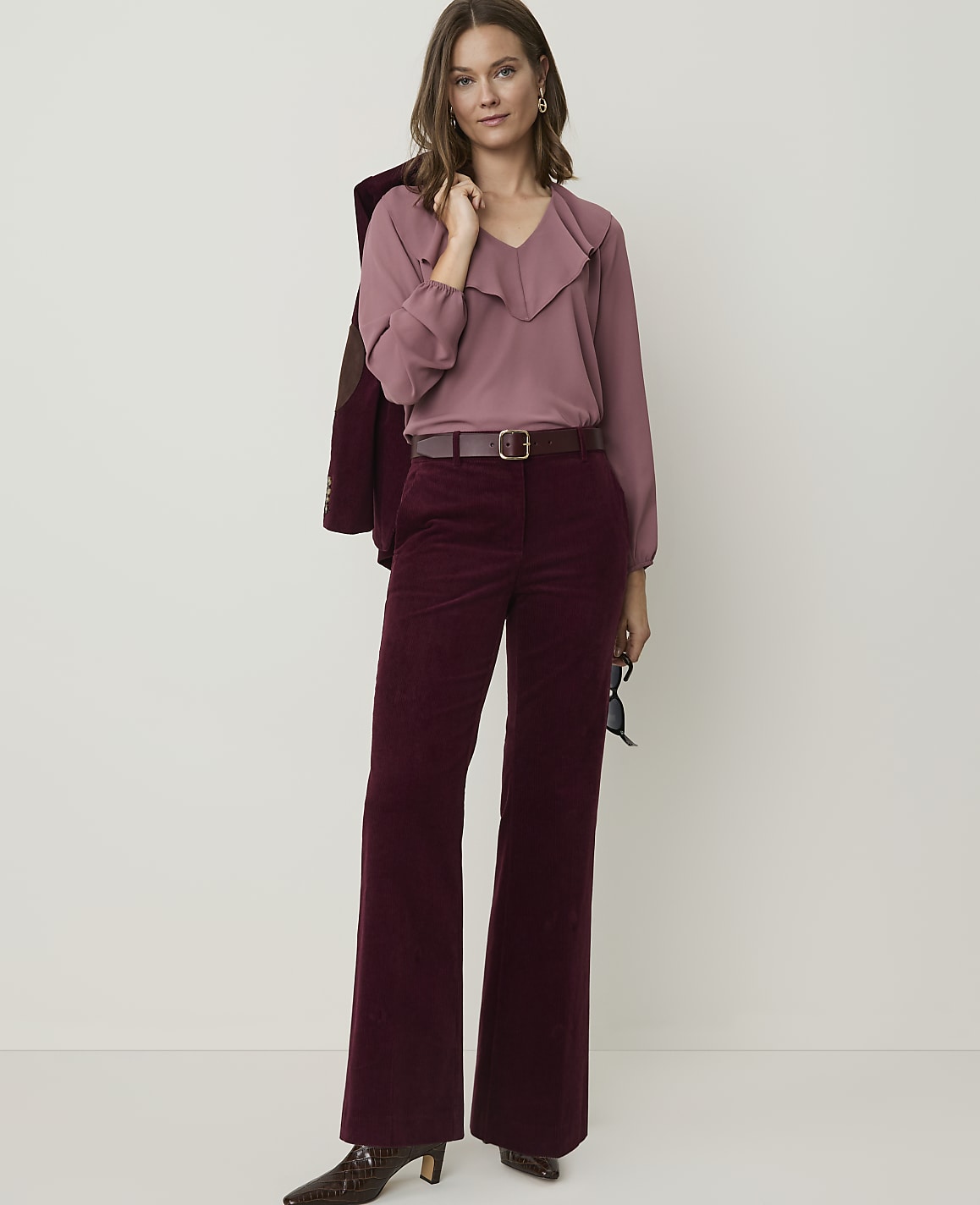 A woman in a mauve ruffled blouse and burgundy corduroy pants stands against a plain, light background, with a maroon jacket draped over her shoulder.