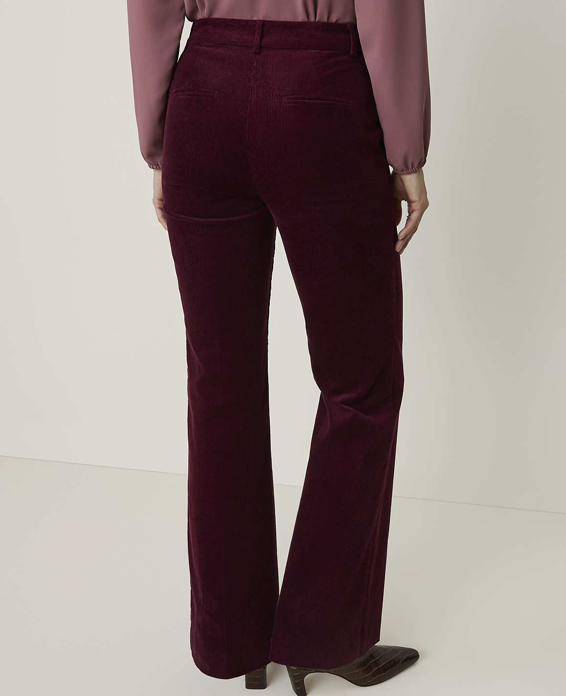 A person wearing burgundy corduroy flared pants and a mauve blouse stands against a plain white background.