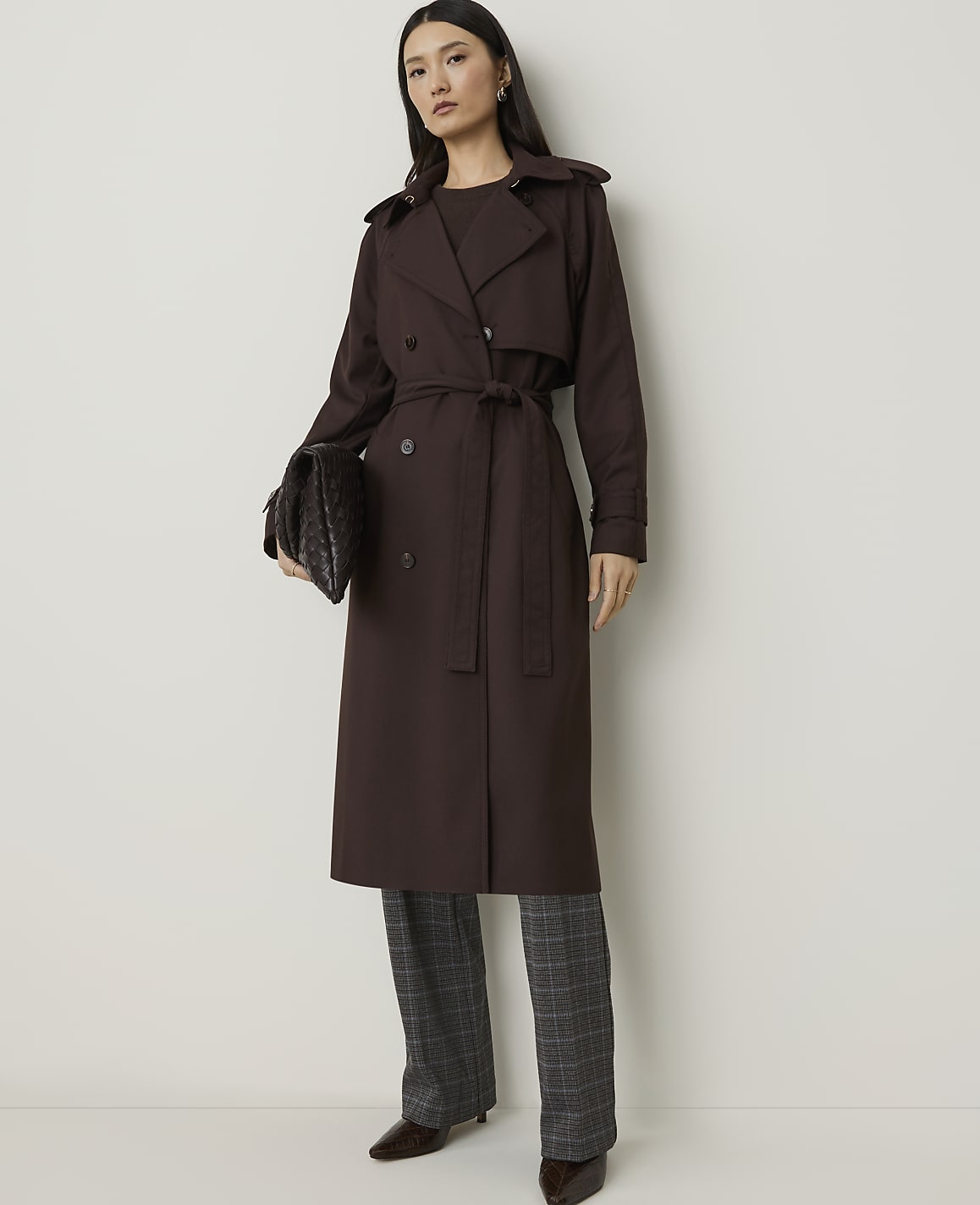 A woman in a brown trench coat and plaid pants stands against a plain white background, holding a dark clutch.