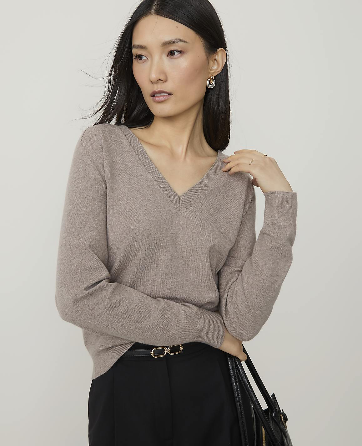 A woman in a taupe v-neck sweater and black pants stands against a plain gray background.