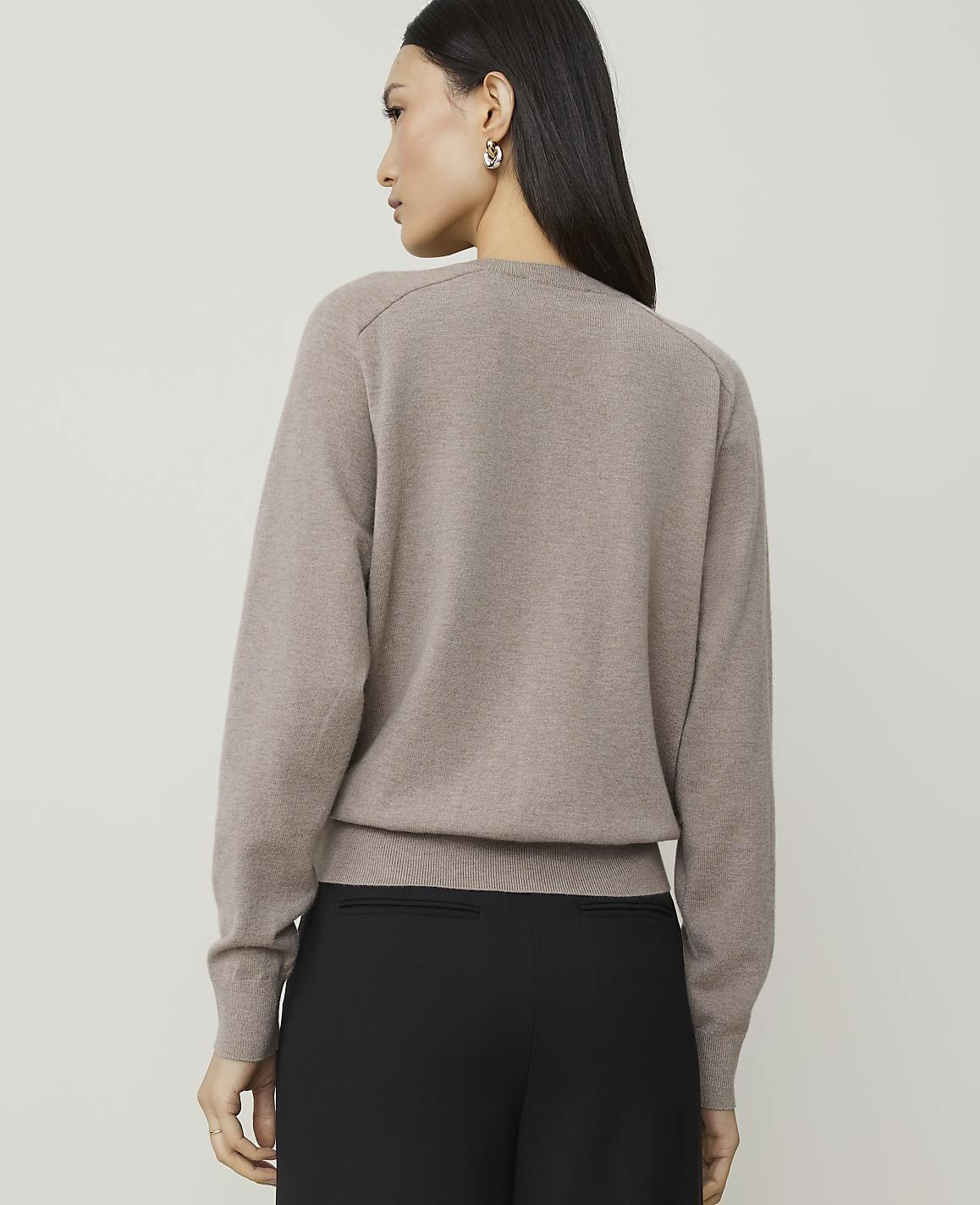 A woman in a taupe sweater and black pants stands against a plain, light gray background.
