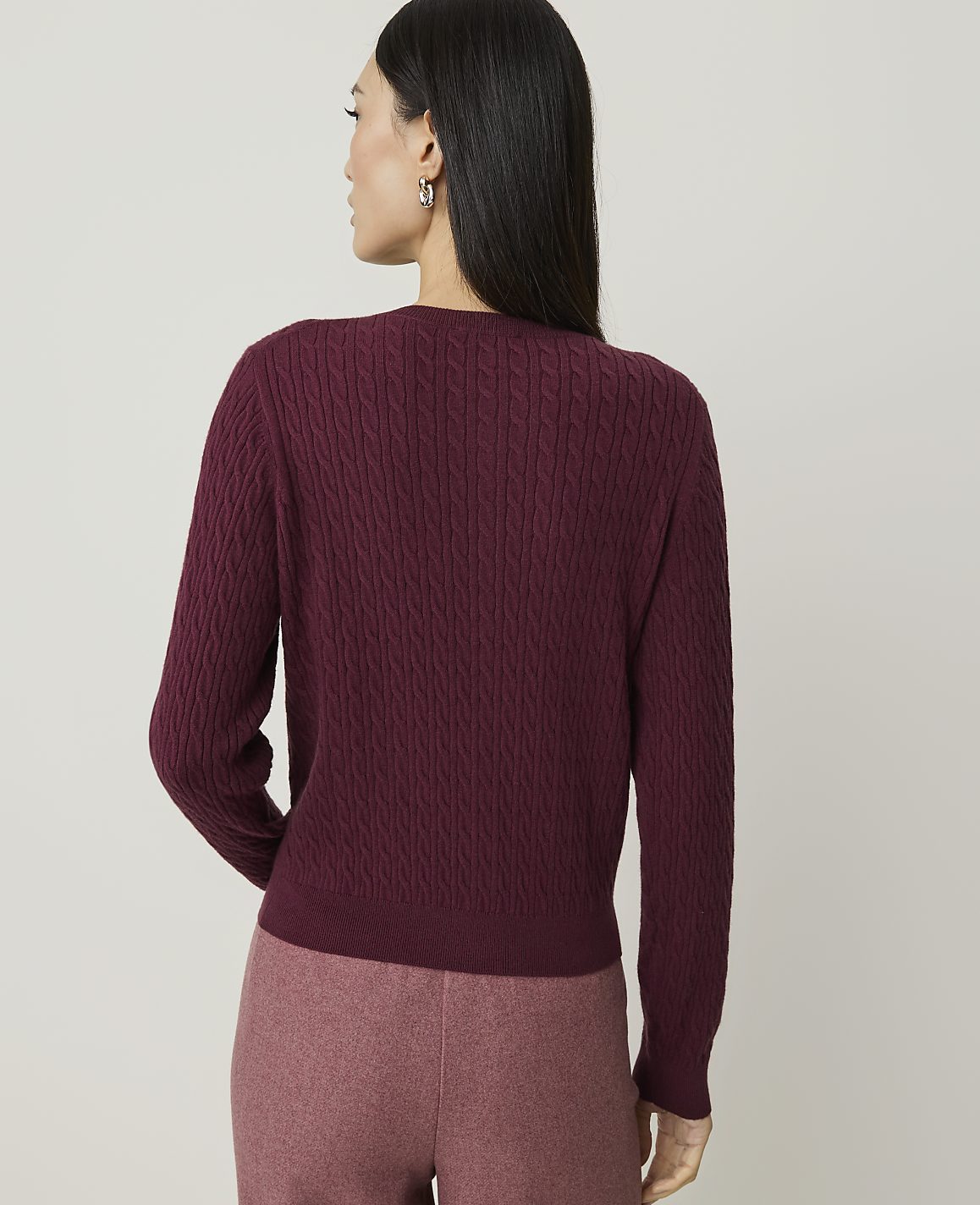A woman in a maroon cable-knit sweater and pink pants stands against a plain, light-colored background.
