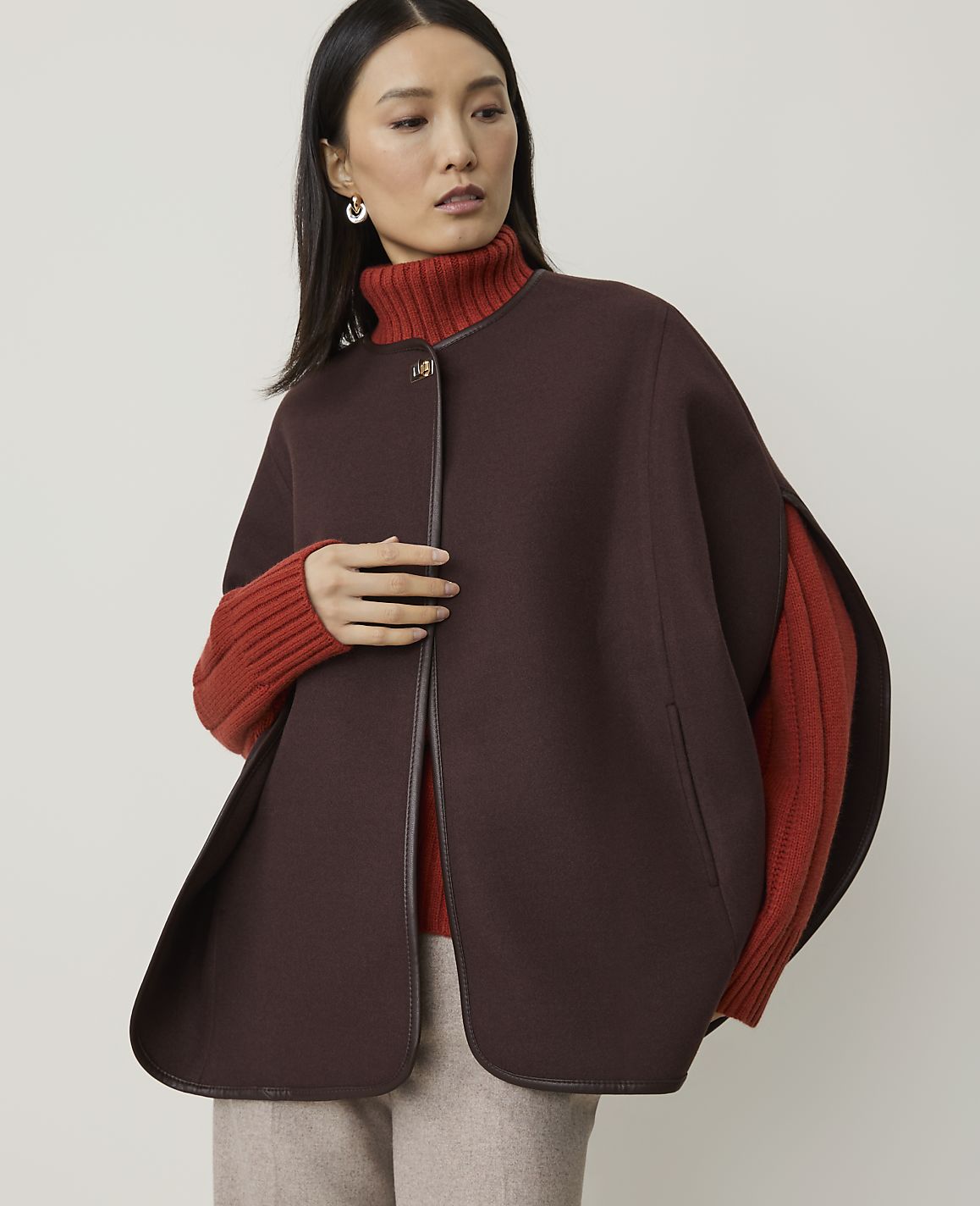 A woman in a brown cape and orange turtleneck stands against a plain, light background.