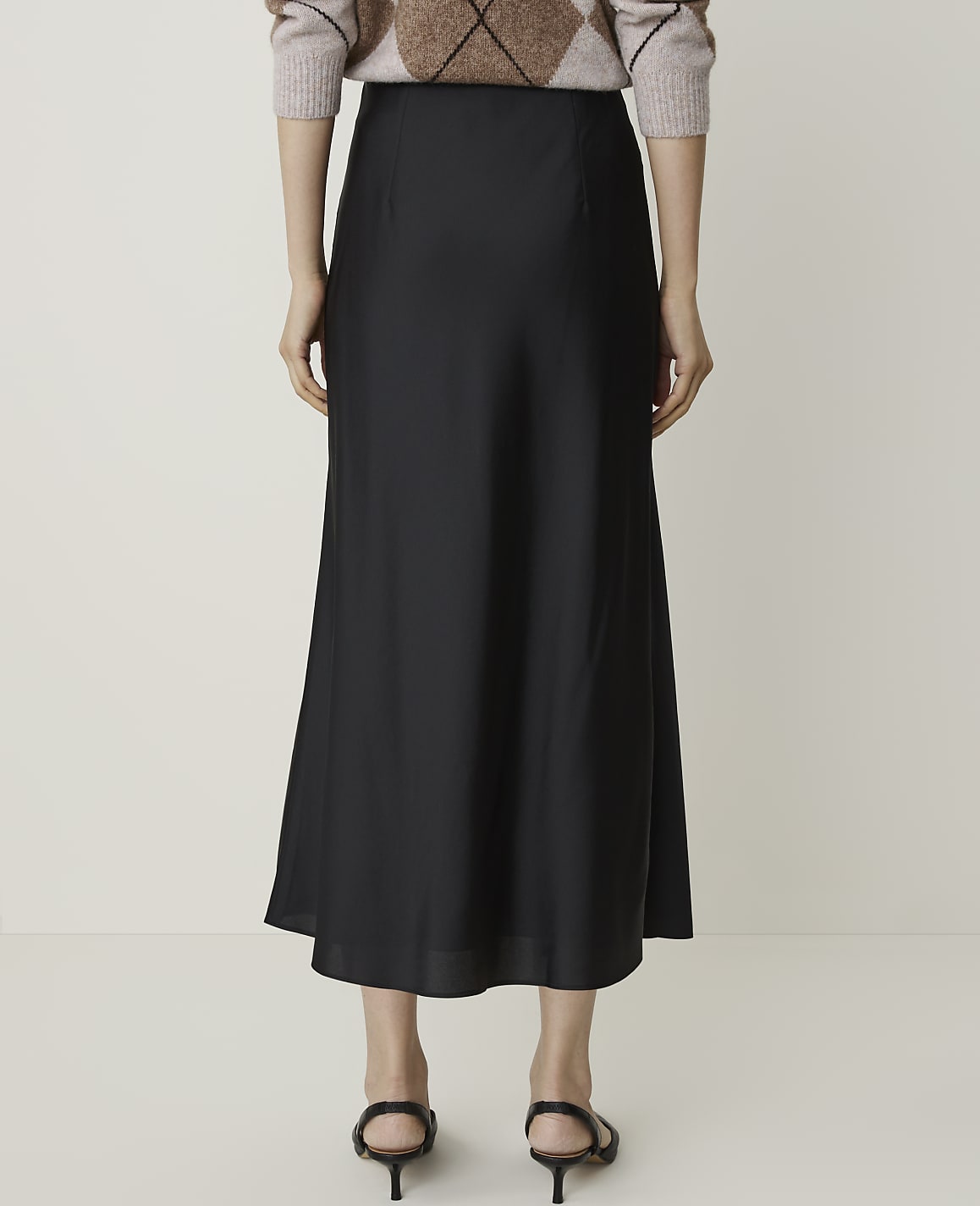 A person stands in a plain studio, wearing a black midi skirt and a patterned sweater, with their back to the camera.